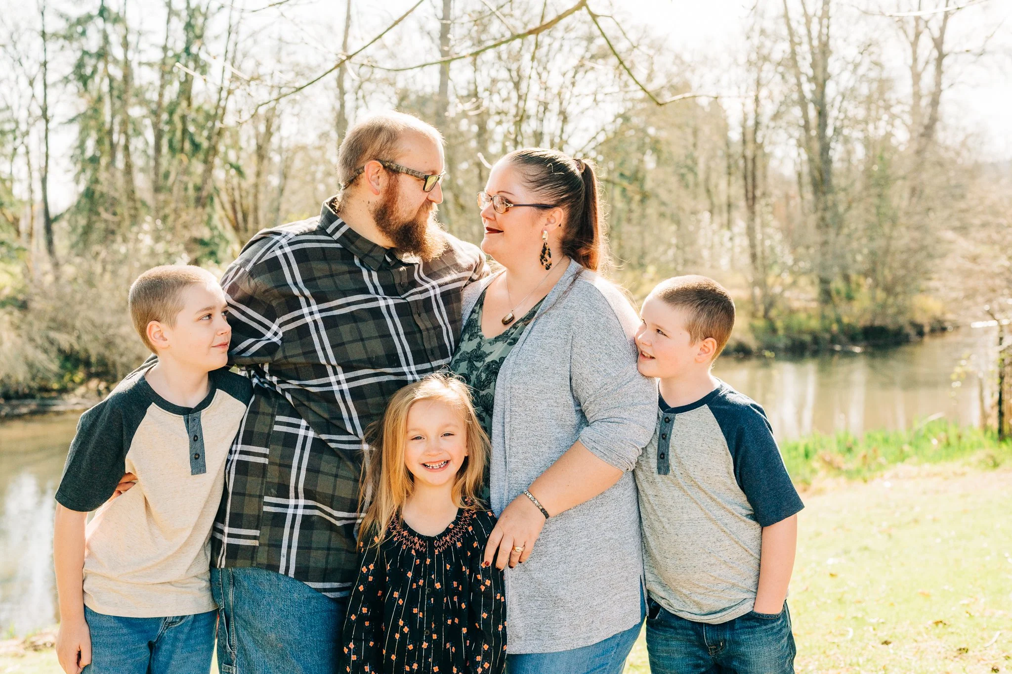 A family of six, including two adults and four children, standing outdoors near a river or lake with trees in the background during daytime. They are smiling and looking at each other, enjoying a sunny day.