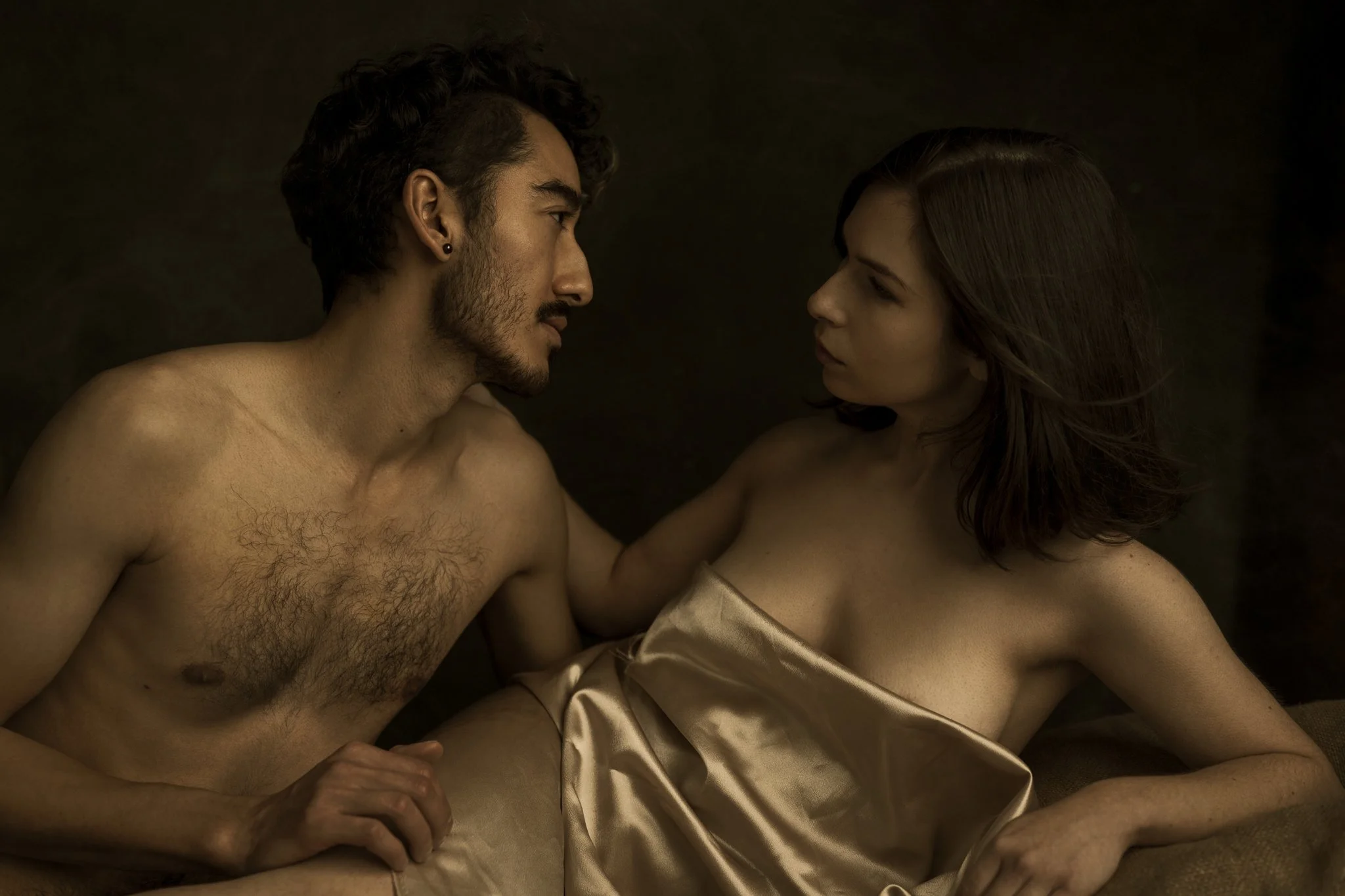 A shirtless man and a woman with a satin sheet covering her torso, sitting close and looking into each other's eyes against a dark background.