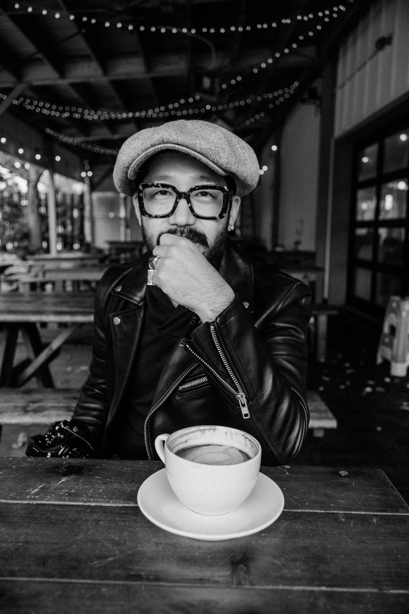 A man with glasses and a beard wearing a flat cap and a leather jacket, sitting at a table with a cup of coffee or tea in front of him, in a rustic, decorated indoor setting.