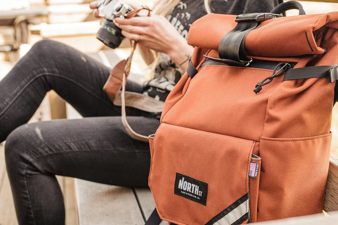 Person sitting on a bench holding a camera, with an orange backpack placed next to them.