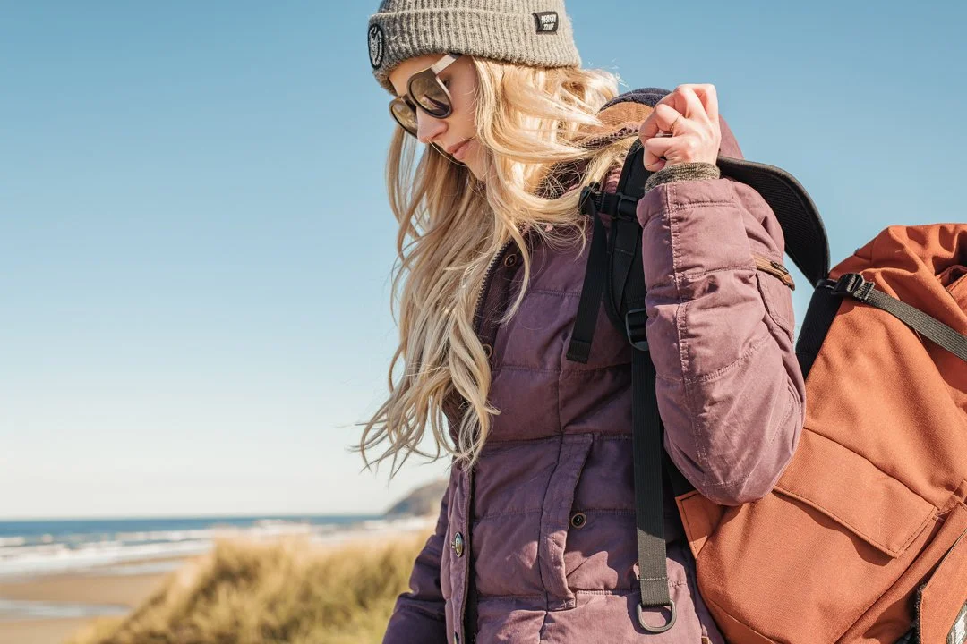 A woman with long blonde hair wearing sunglasses, a beanie, a purple jacket, and carrying an orange backpack stands outdoors near the beach on a clear day.