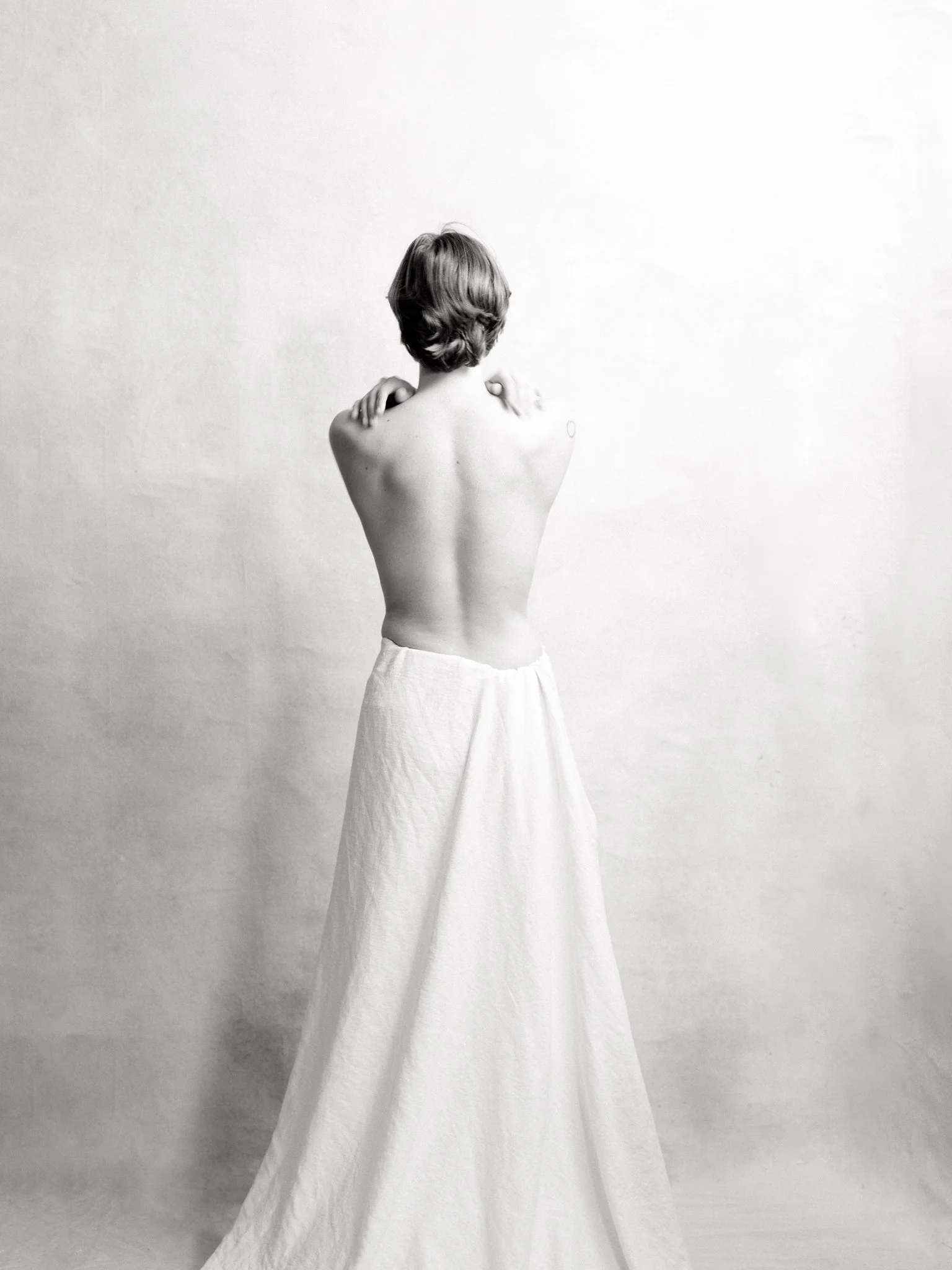 Back view of a topless woman with short hair, wearing a long white skirt, standing against a plain light-colored background.
