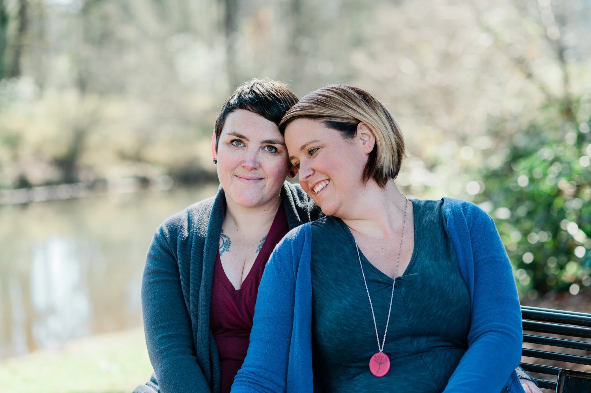 Two women sitting closely outdoors near a body of water, sharing a tender moment with one leaning her head on the other's shoulder, smiling.