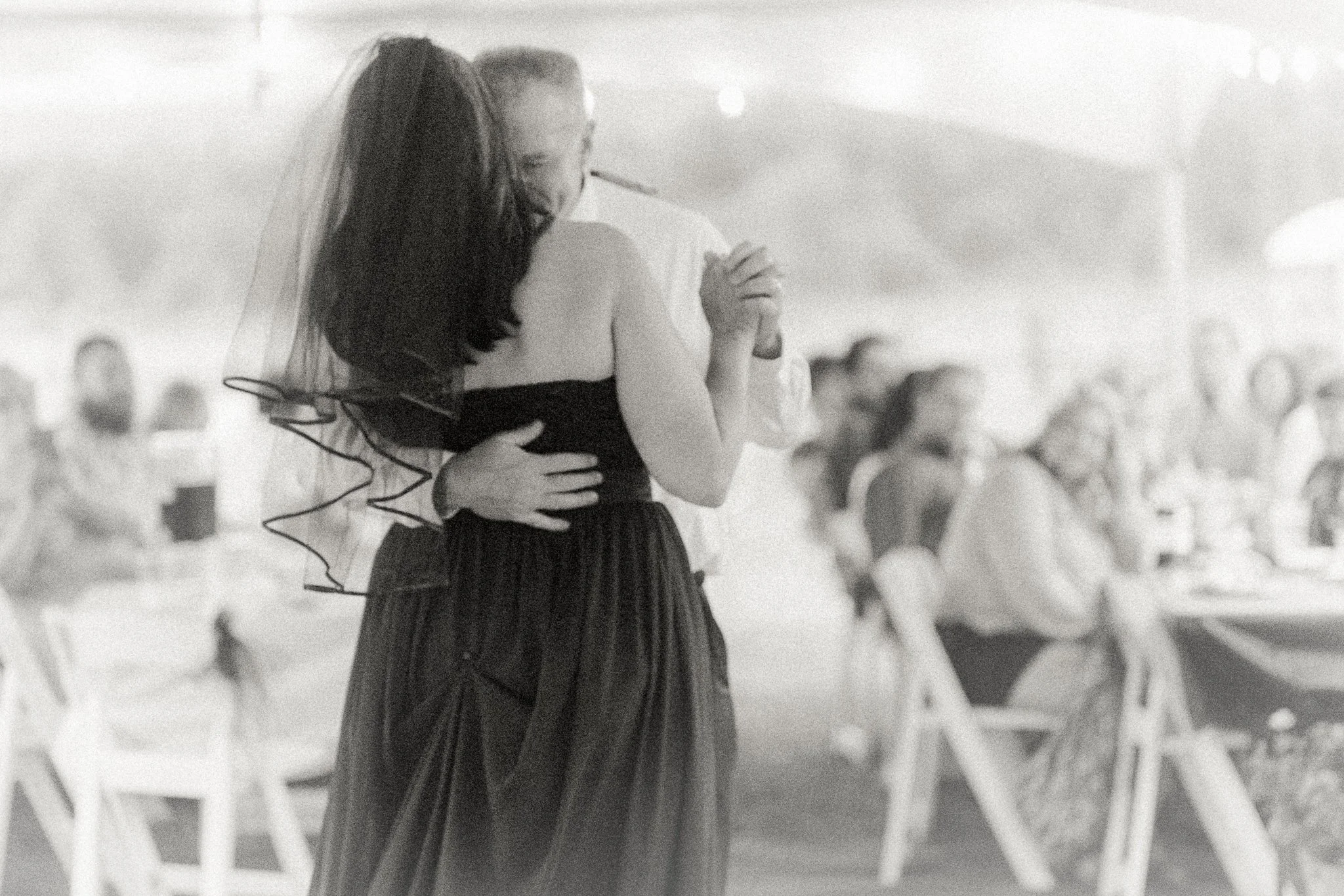 A bride and groom dance closely, holding each other, at their wedding reception, with guests sitting at tables watching them.
