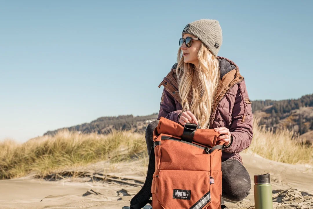Woman wearing sunglasses and a beanie with a backpack on sandy terrain with grass and hills in the background.