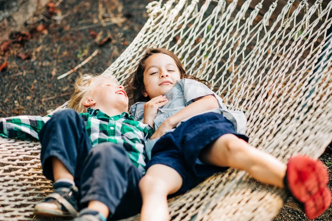 Two children lying together on a hammock outdoors, smiling and relaxed.