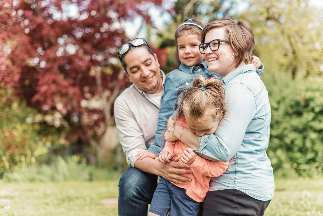 A family of four smiling and hugging outdoors in a park during fall, with colorful autumn trees in the background.