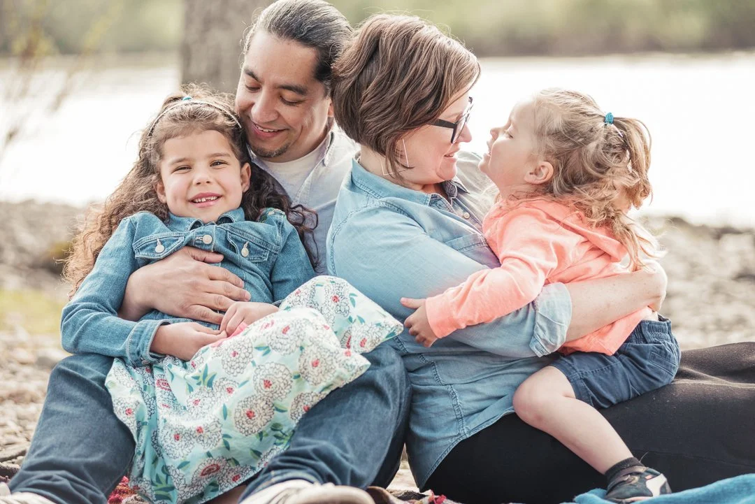A family sitting outdoors by a lake, two women and two young girls, hugging and smiling.