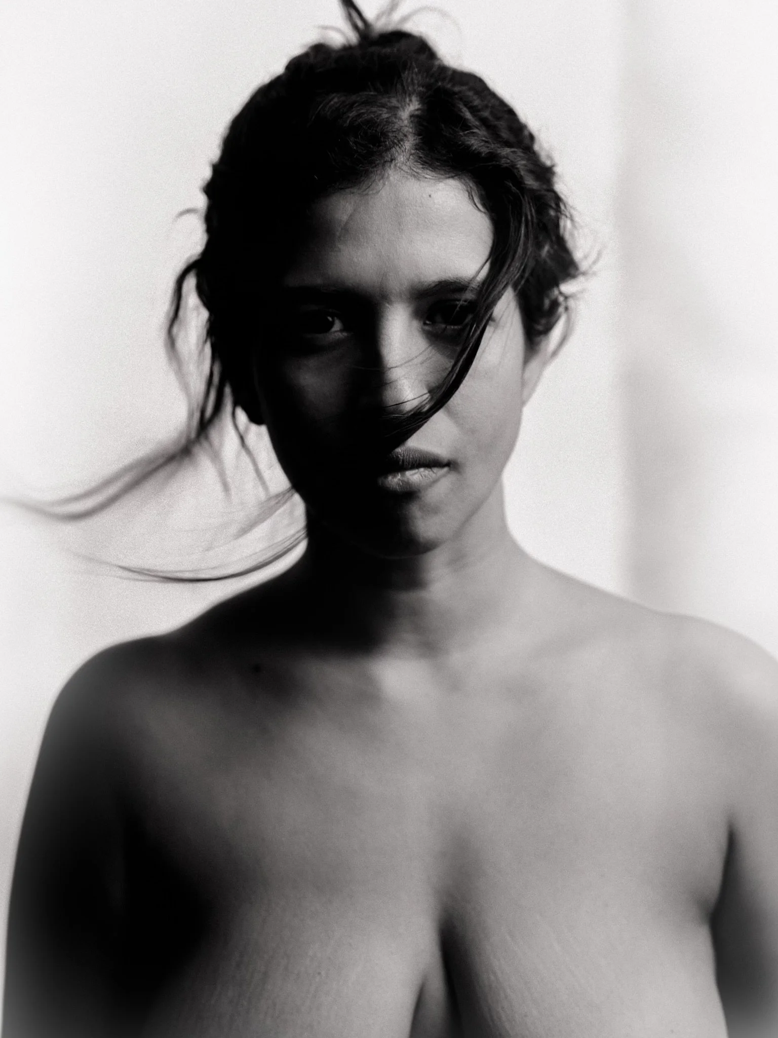 Black and white portrait of a topless woman with dark hair, partially covering her face, looking directly at the camera.