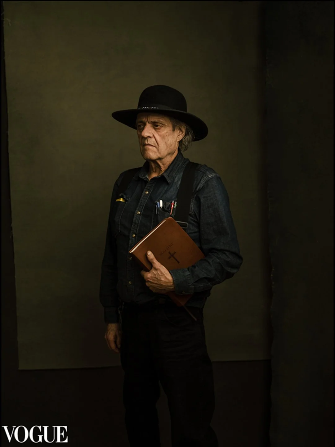 An elderly man wearing a black hat and dark clothing, holding a Bible with a cross on the cover, standing against a plain background, Vogue magazine.