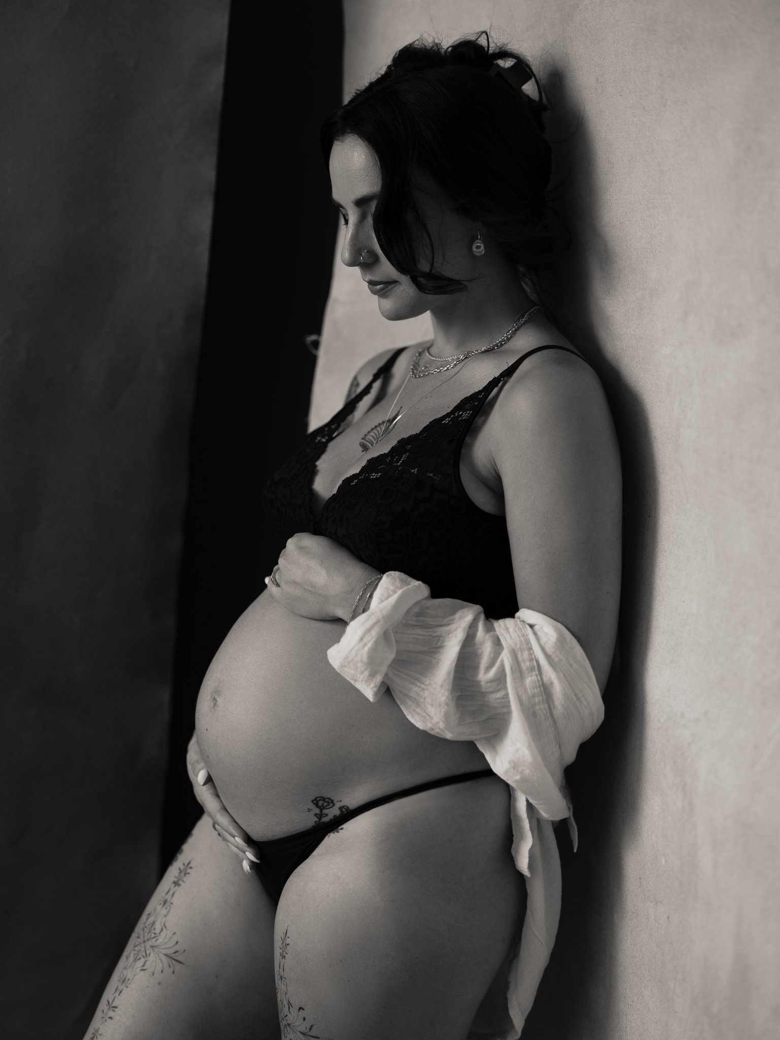 A pregnant woman with dark hair, dressed in black lingerie, standing against a wall with her eyes closed, holding her belly.