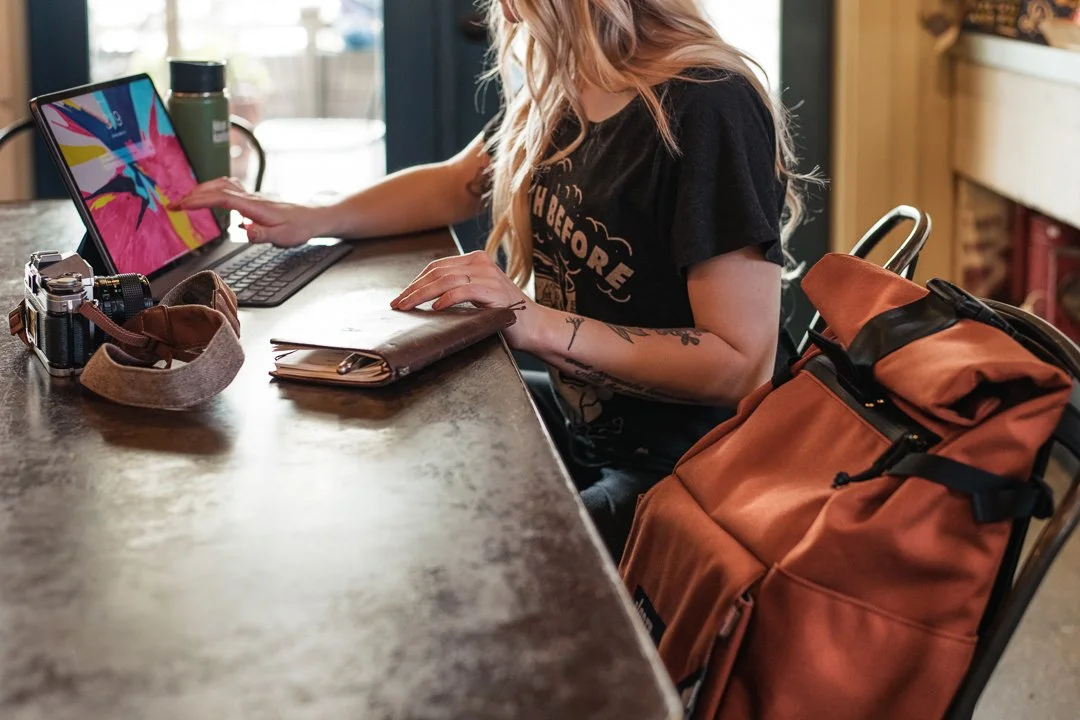 A woman with long, wavy blonde hair sitting at a wooden table, using a tablet with a colorful abstract wallpaper, with a brown handbag and a camera next to her. Sunlight coming through a window in the background.