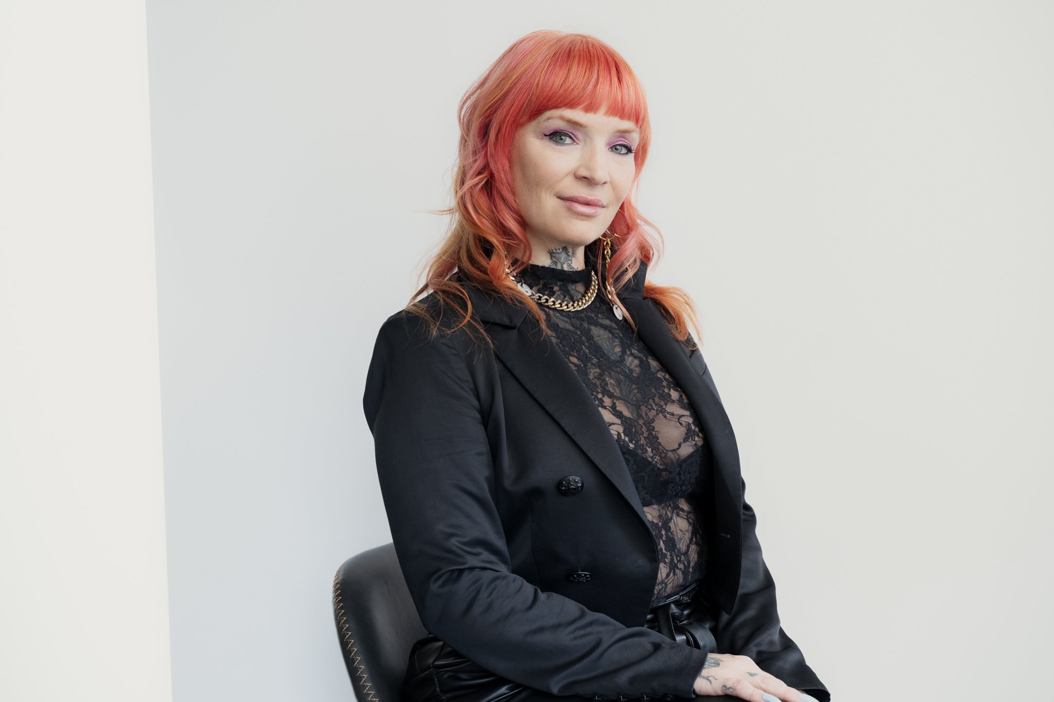 woman with red hair wearing black lace top and blazer, sitting on a black chair against a white wall.