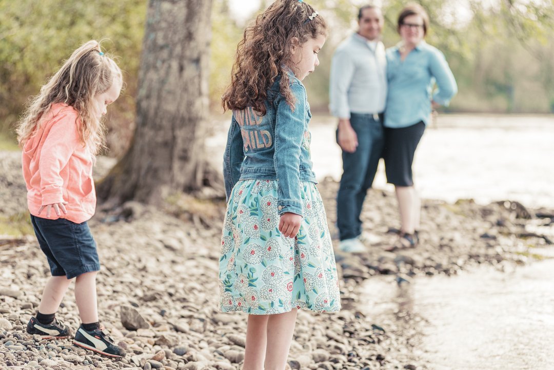 A family of four standing by a lake in a natural outdoor setting, with two young girls in the foreground and a man and woman in the background.