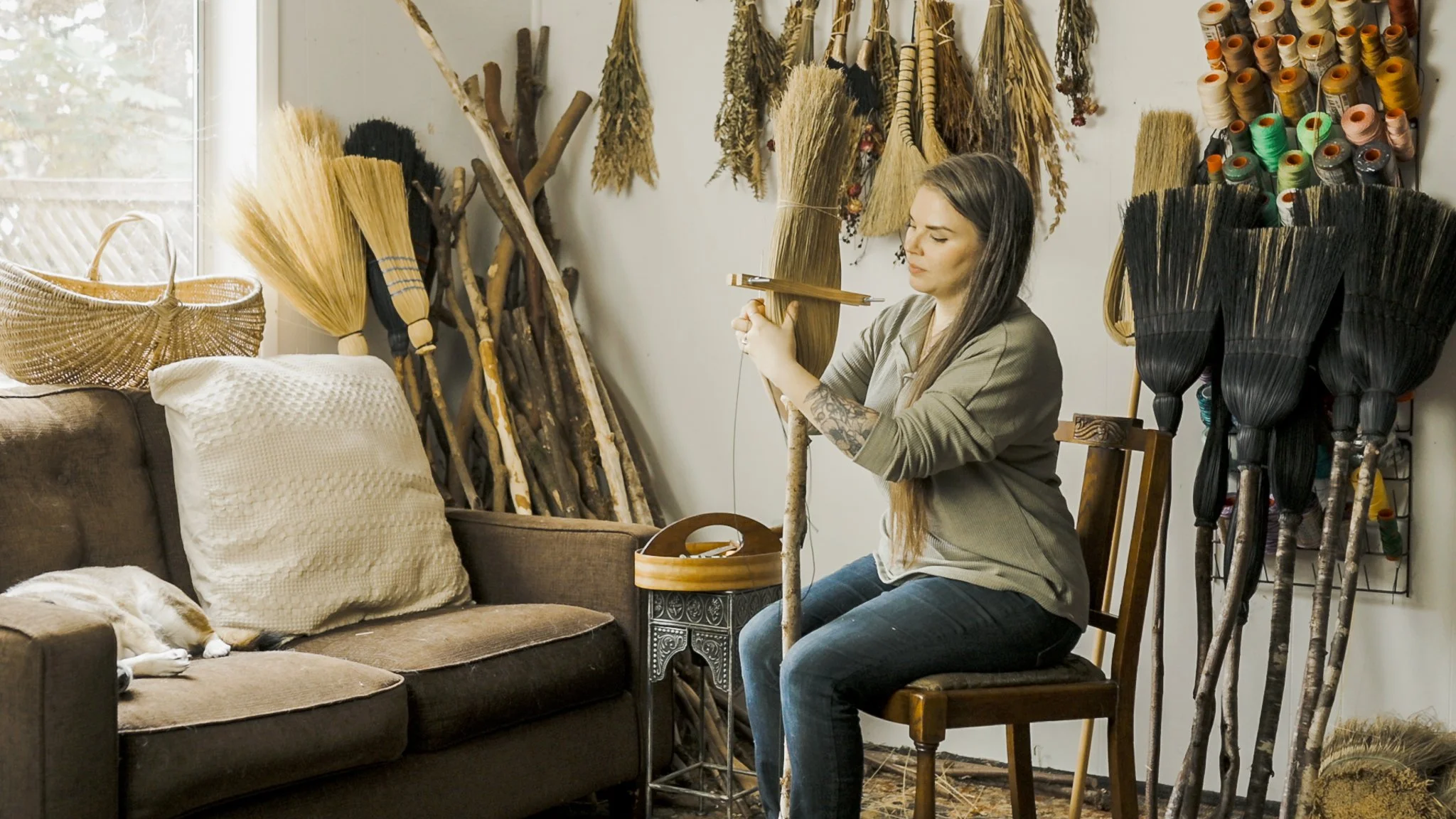 A woman sitting on a wooden chair with a tattoo on her right forearm, weaving a willow branch into a basket. There are various dried plants and broomsticks hanging on the wall and resting against the wall, with a dog sleeping on a brown couch nearby 