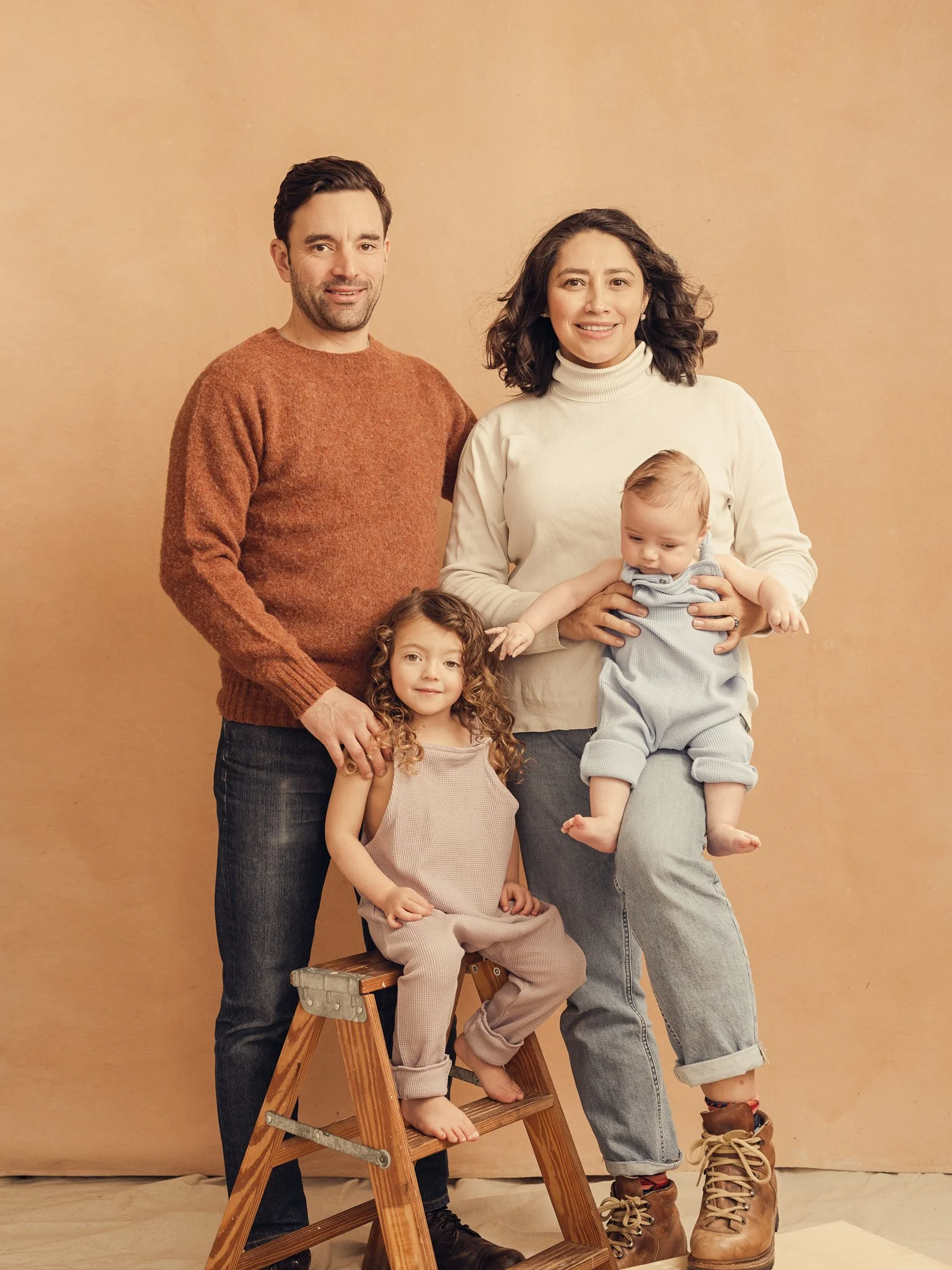 A family of four posing against a beige background, including a man, a woman holding a baby, and a girl sitting on a wooden step stool.