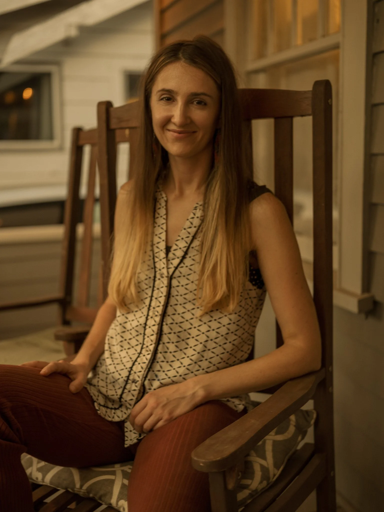 A woman with long, light brown hair sitting on a wooden rocking chair on a porch at night, smiling at the camera.
