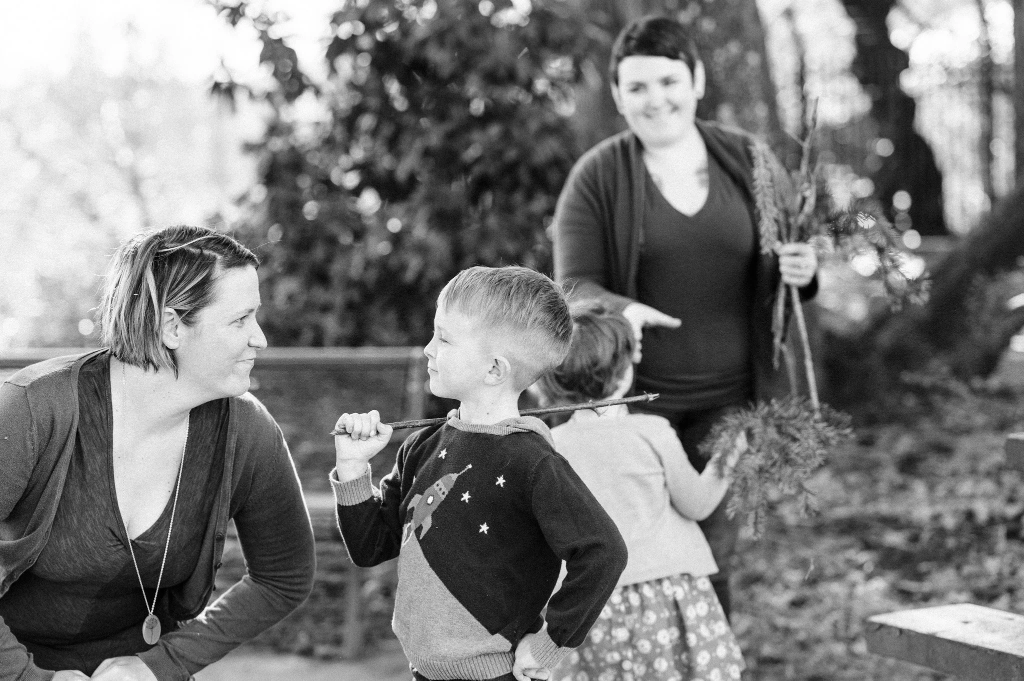 A black and white photo of a woman and three children outdoors, with trees in the background. The woman is leaning forward, smiling at a young boy who is holding a stick on his shoulder. In the background, a woman is holding a bundle of branches, and