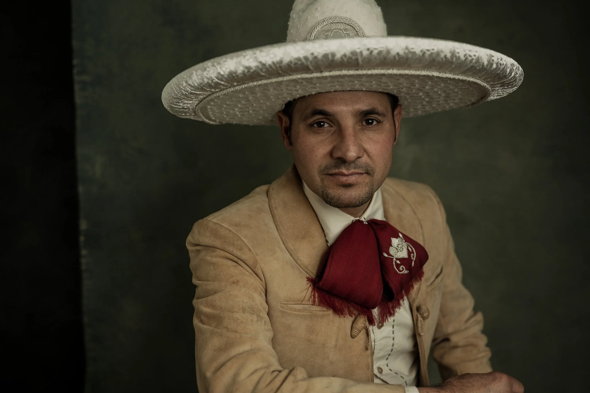 A man wearing traditional Mexican attire with a large sombrero, a tan jacket, a white shirt, and a red neckerchief.