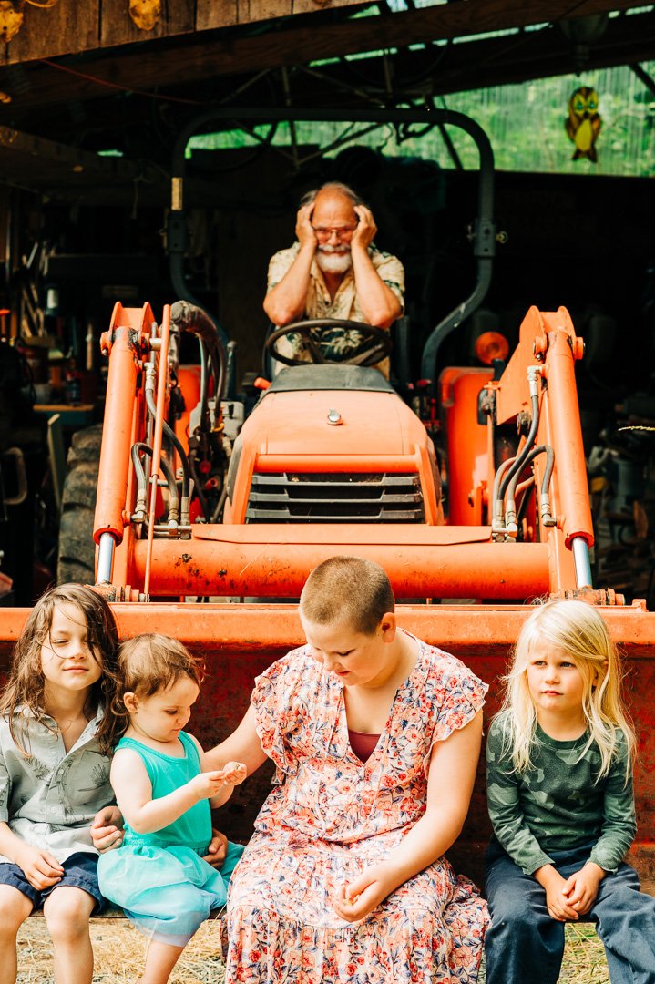 An elderly man with glasses and a beard is sitting on a tractor, smiling and holding his head with both hands, while four children sit in front of him, on the ground, outdoors.