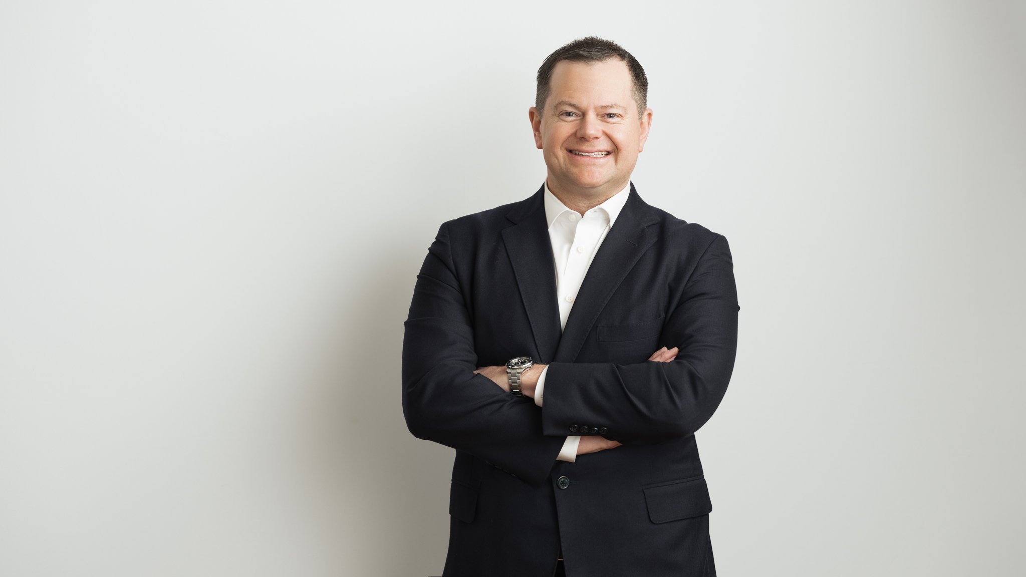 A smiling man with short dark hair wearing a black suit, white dress shirt, and watch, standing with arms crossed against a plain light-colored background.