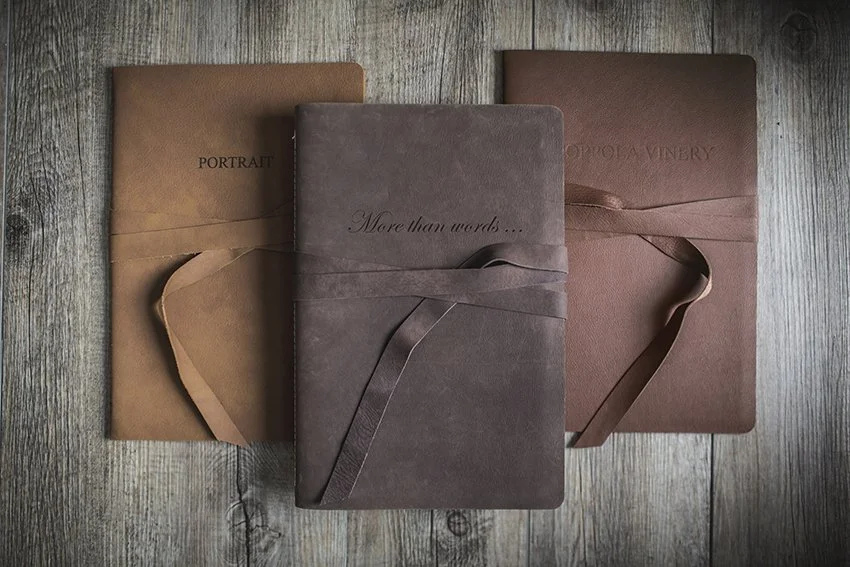 Three brown book covers with ribbon closures, placed on a wooden surface. The titles are 'PORTRAIT,' 'More than words...', and 'OOPOLA VINEY.'
