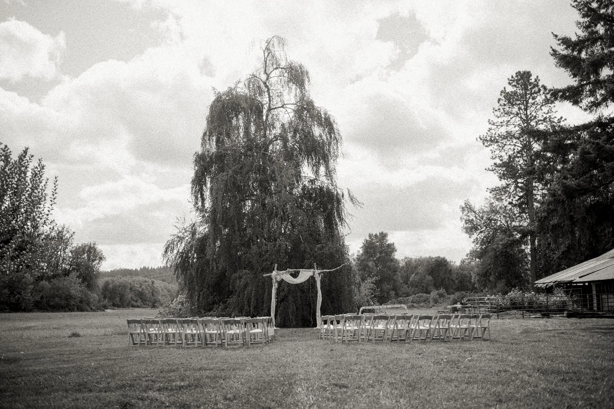 Outdoor wedding setup with chairs arranged in front of an arch made of wood and fabric, in a grassy field with trees and a cloudy sky.