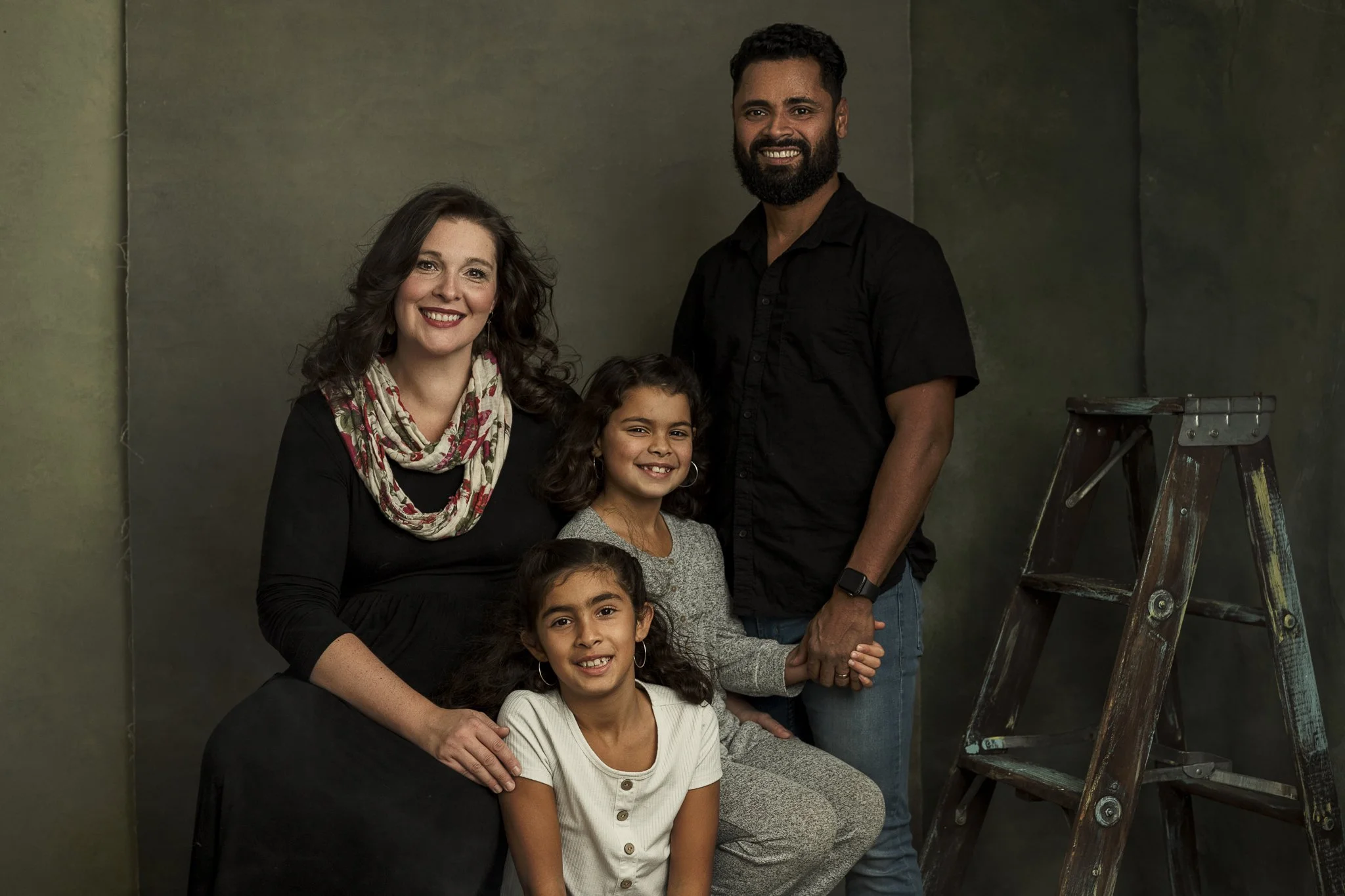 A family portrait of four people, including a woman, a man, and two young girls, posing indoors with a greenish background and a wooden ladder.