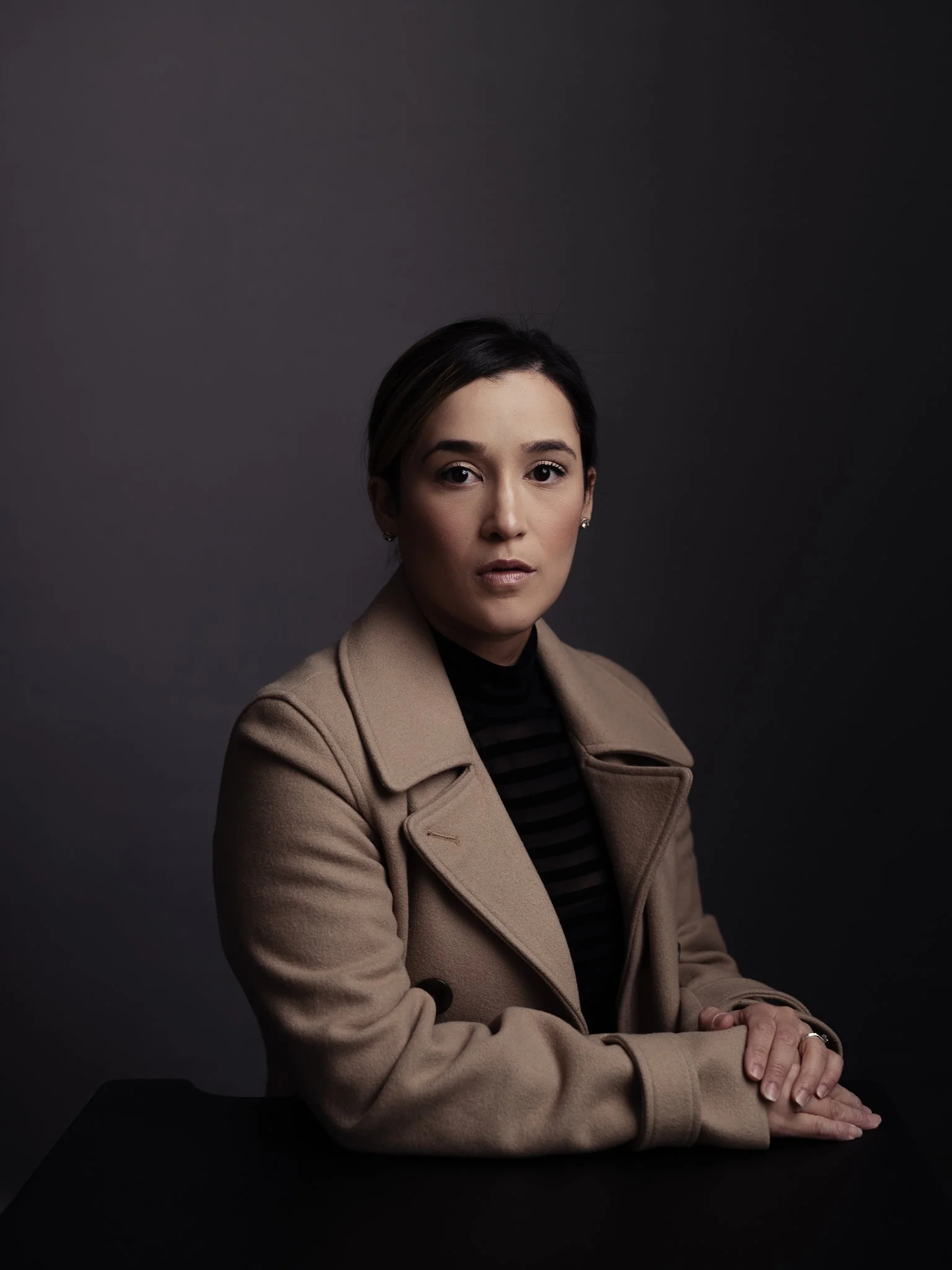 Portrait of a woman with black hair, wearing a tan coat and black striped top, sitting at a black table against a dark gray background.