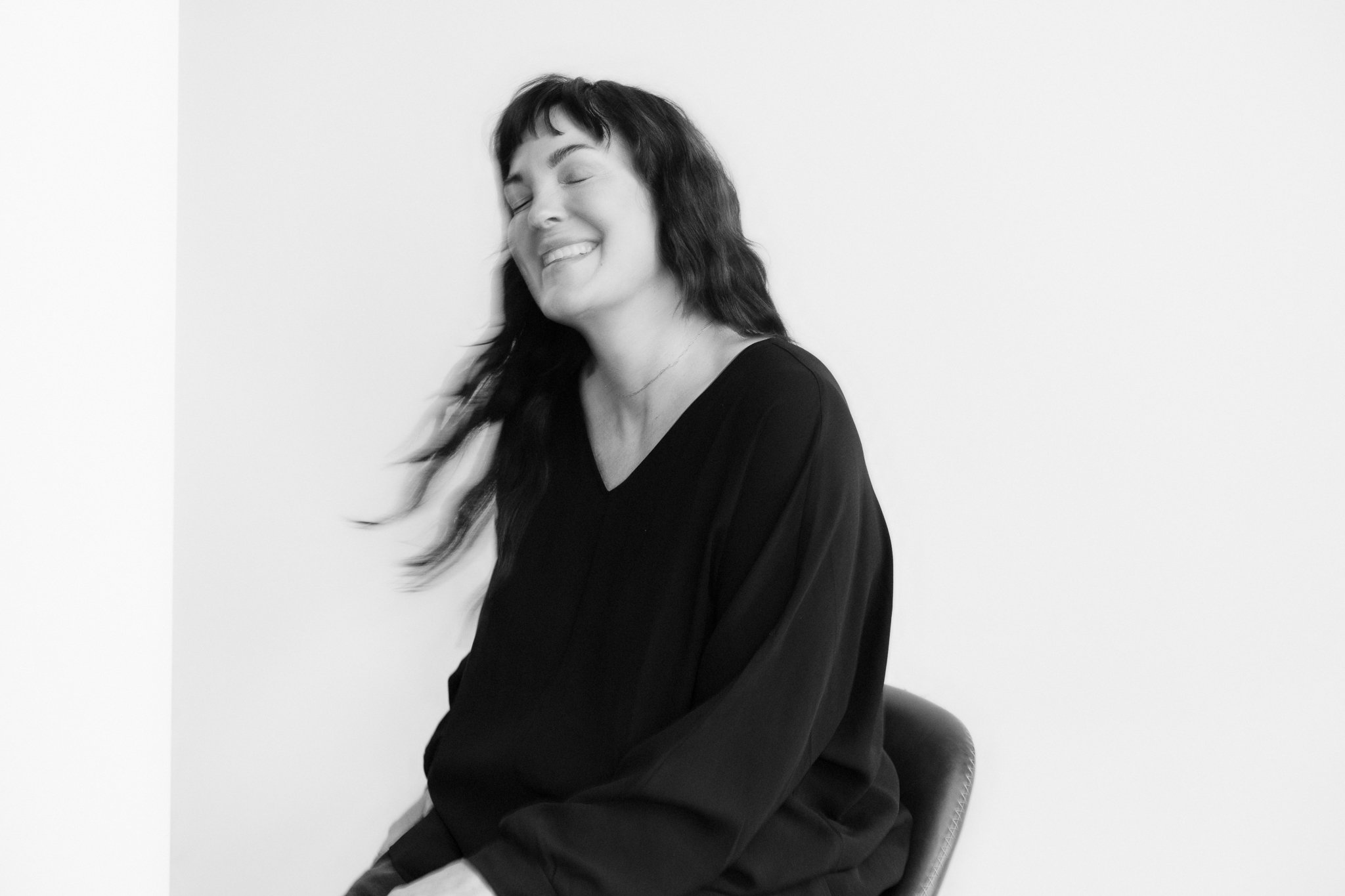 A woman with dark hair and bangs smiles with her eyes closed, sitting on a chair against a plain white wall.
