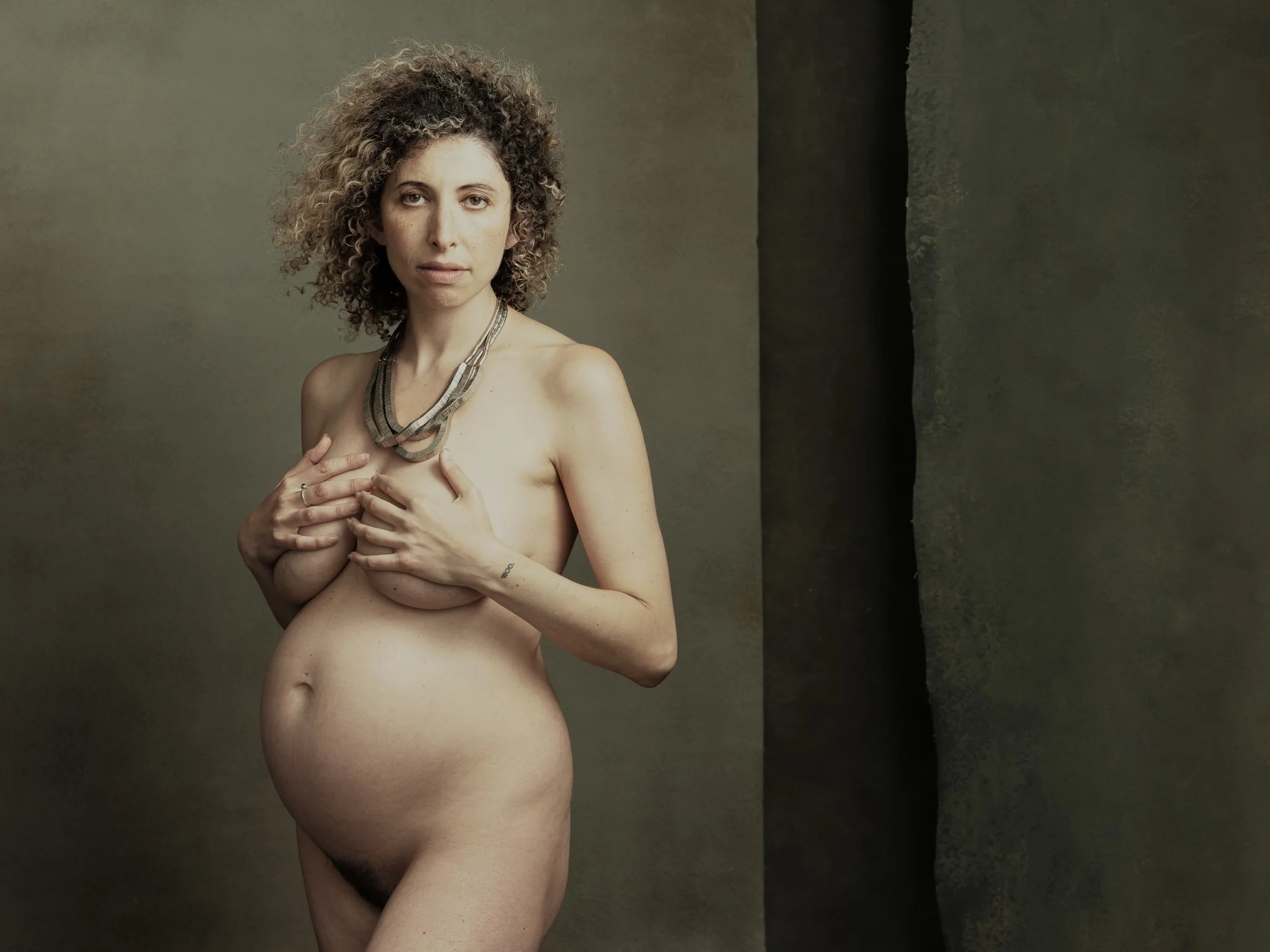 A nude woman with curly hair and a necklace, standing against a textured background, covering her chest with her hands.