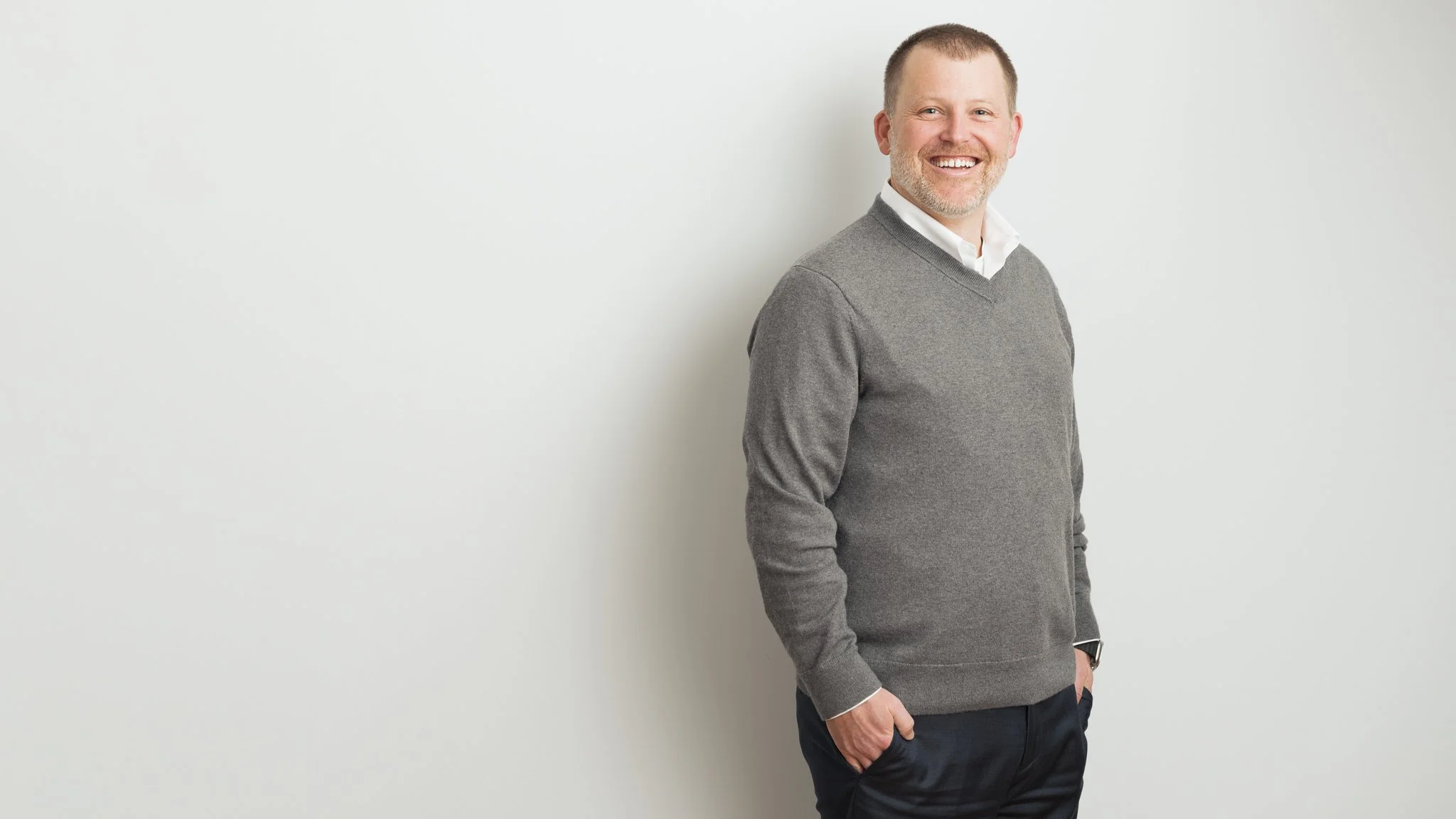 A smiling man with light hair and a beard, wearing a gray sweater over a white collared shirt and dark pants, standing against a plain white wall.
