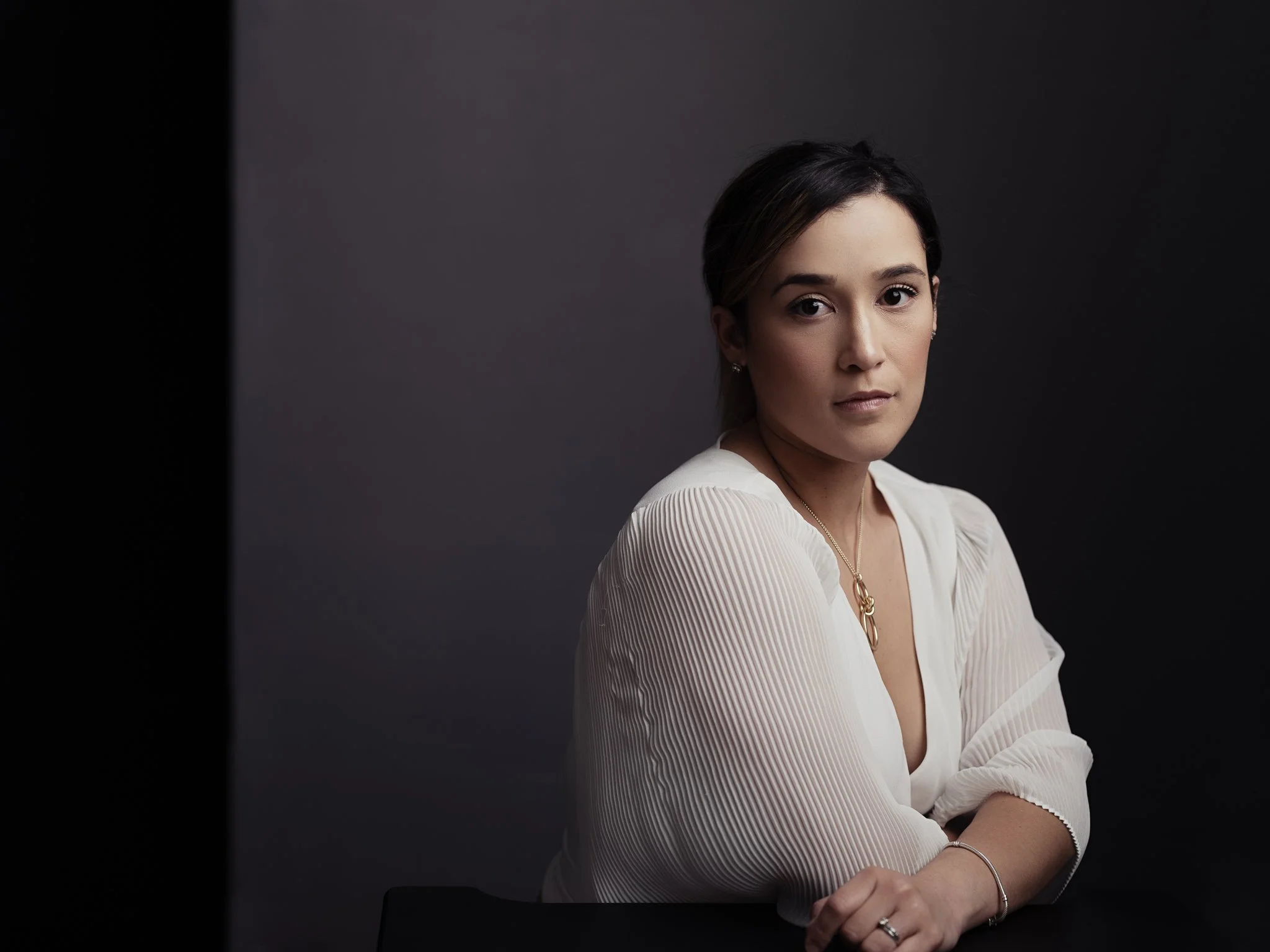 Professional headshot of a woman with dark hair, light skin, and brown eyes looking at the camera, wearing a white pleated blouse, a gold necklace, and a bracelet, sitting with folded arms against a dark background.