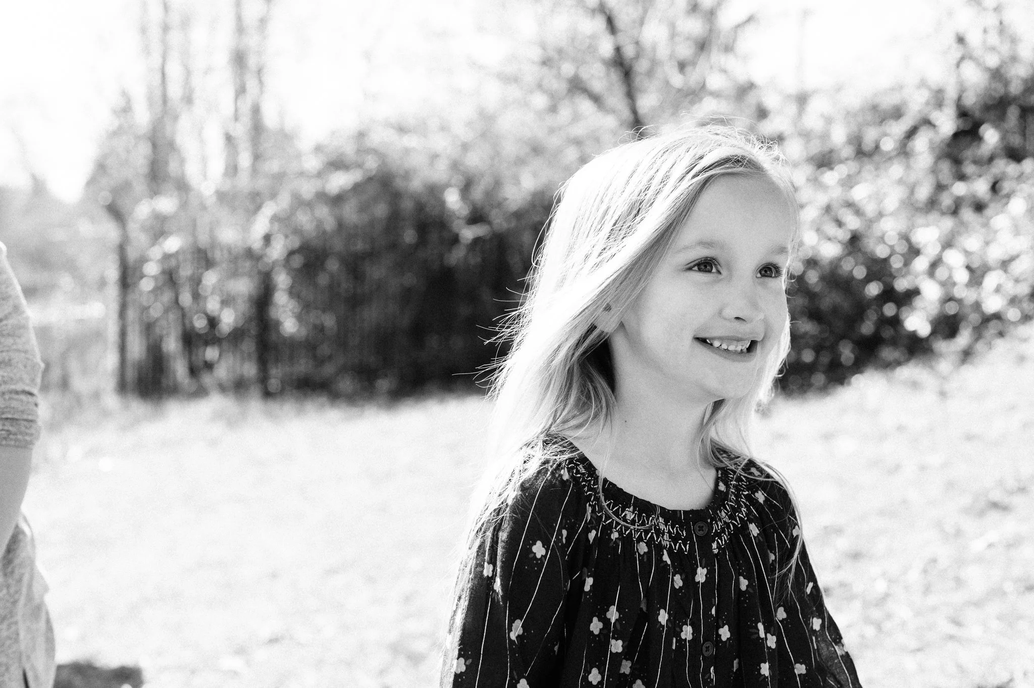 Black and white photo of a smiling young girl with long hair outdoors, wearing a floral patterned dress.