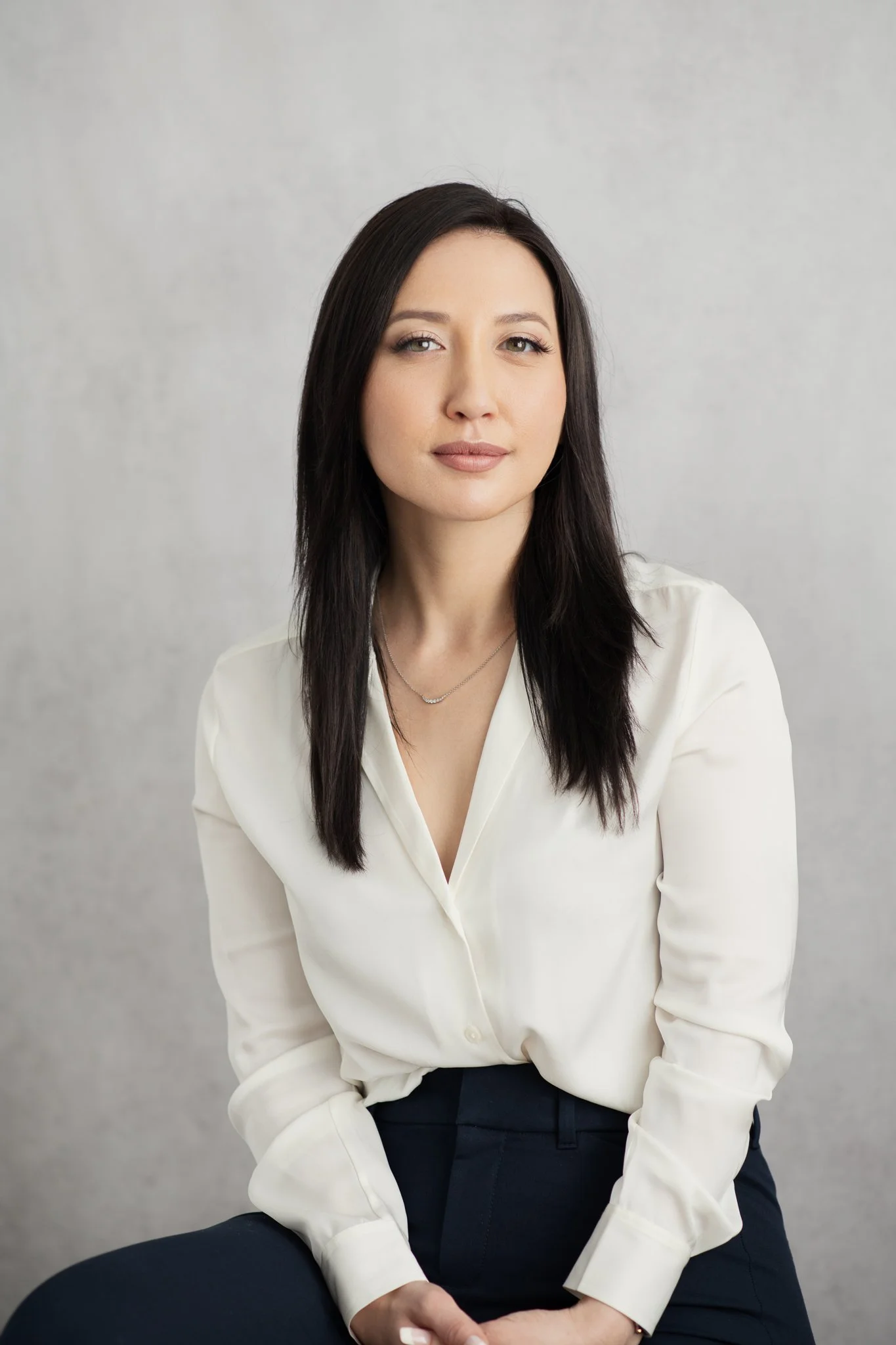 A woman with long dark hair, wearing a white blouse and dark pants, seated against a plain light gray background.