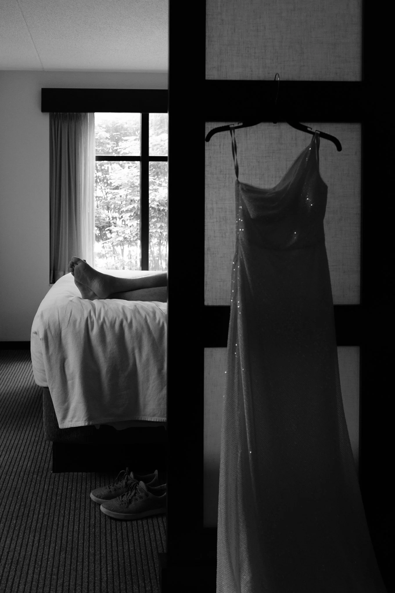 A wedding dress hangs on a door with a hanger, partially obscuring a person lying on a bed in a room with a window and curtains.