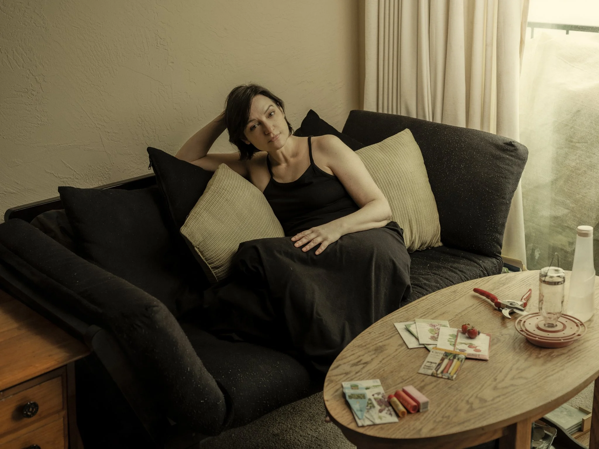 A woman lies on a black sofa surrounded by multiple pillows, with a beige curtain and window in the background. A wooden table with gardening tools, seed packets, and some strawberries is in the foreground.