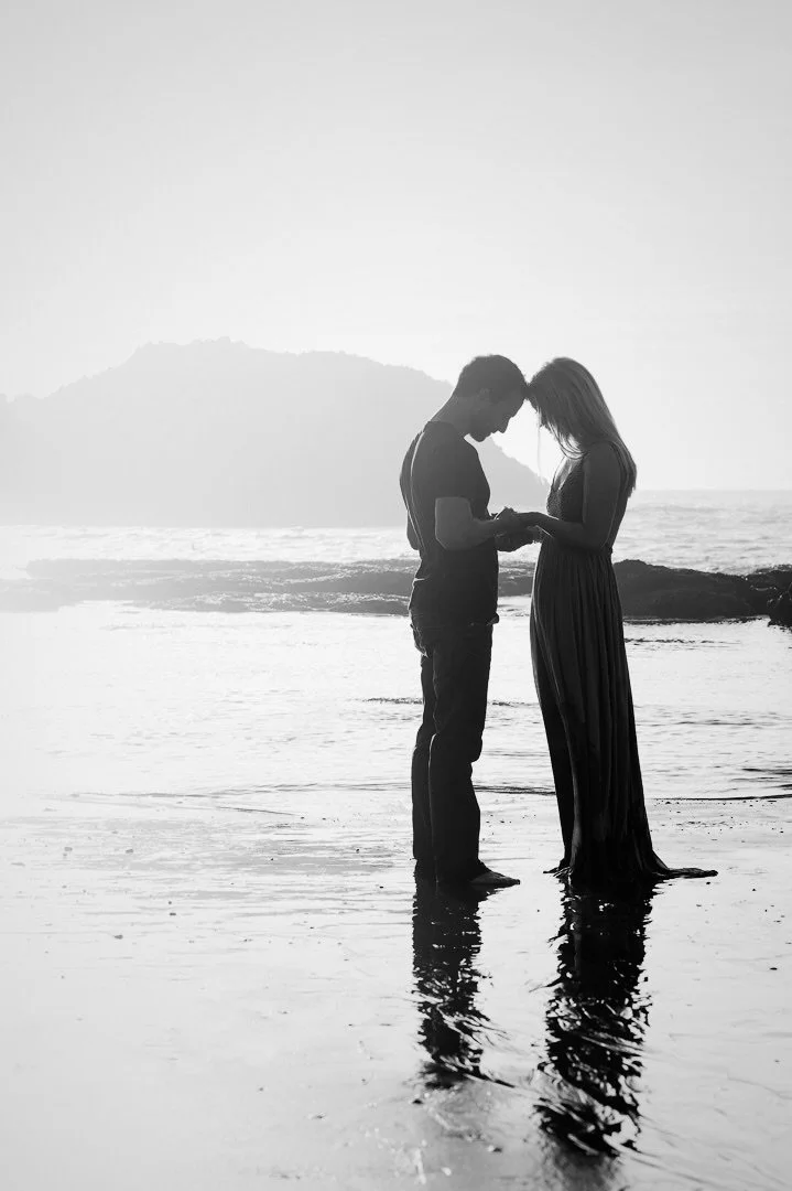 A silhouette of a couple standing close together on a beach, holding hands, with an island in the background, during sunset or sunrise.