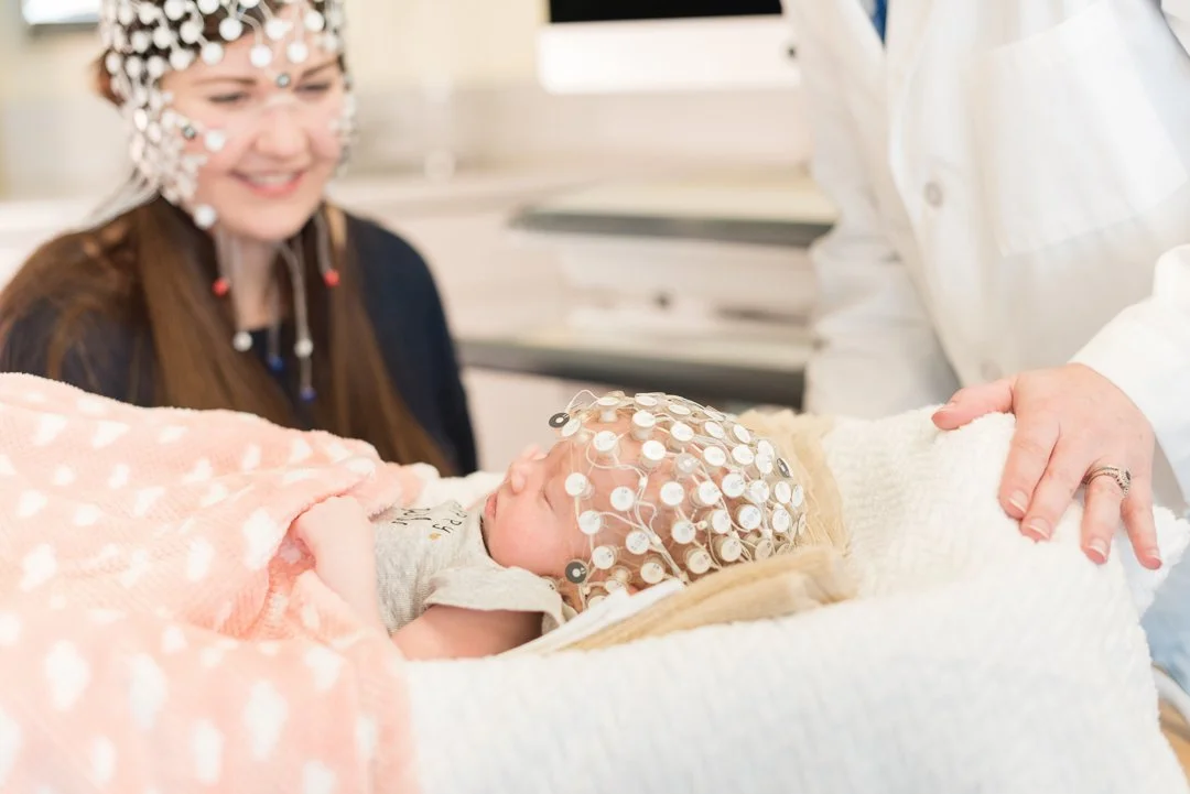 Newborn baby with EEG electrodes on head lying on hospital bed with a pink blanket, surrounded by smiling woman with decorated cap and medical staff.