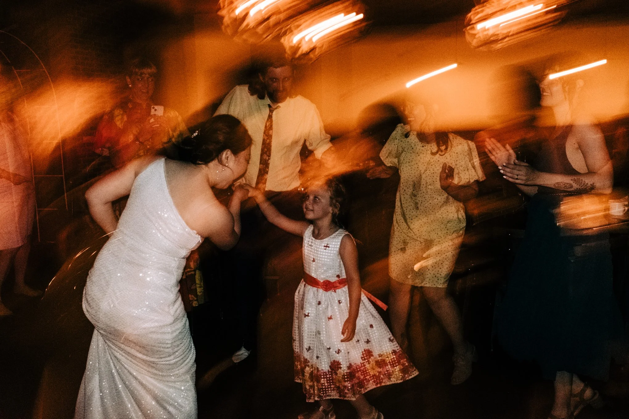People dancing and socializing at a party or celebration, with a young girl in a white dress holding hands with a woman in a white dress, and others in the background.