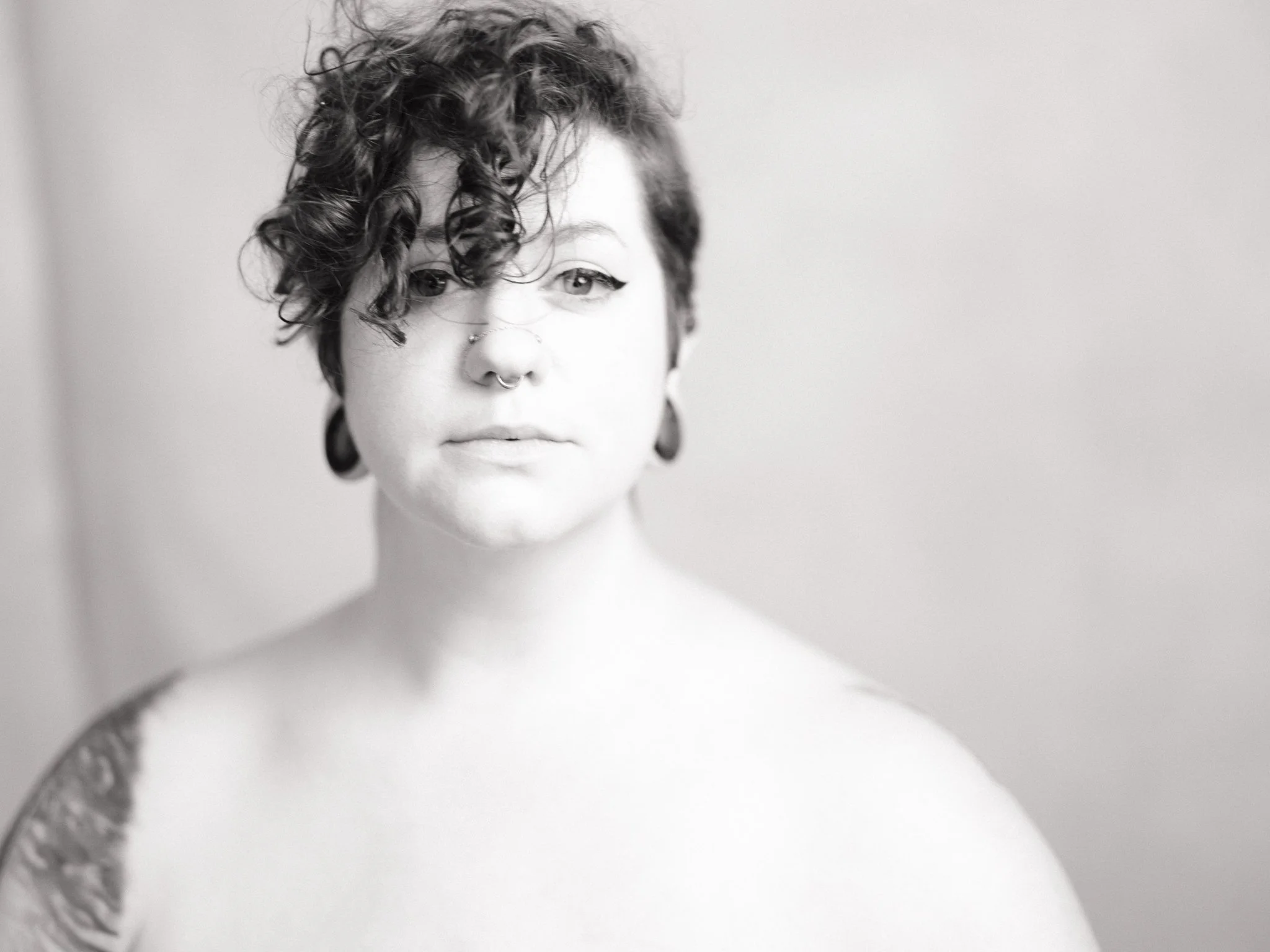Black and white portrait of a woman with short curly hair, septum piercing, and earrings, looking directly at the camera.