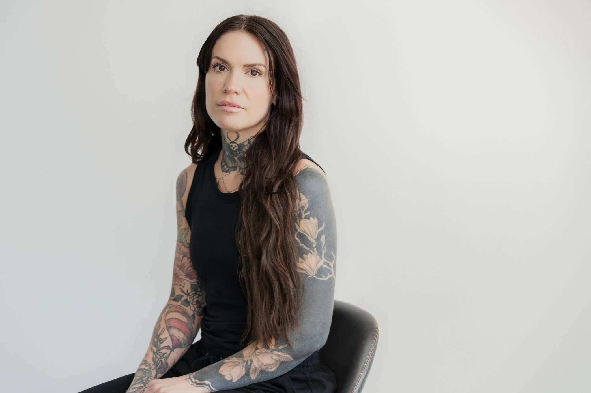 A woman with long wavy brown hair and tattoos on her arms and neck, wearing a black sleeveless top, sitting on a chair against a plain white background.