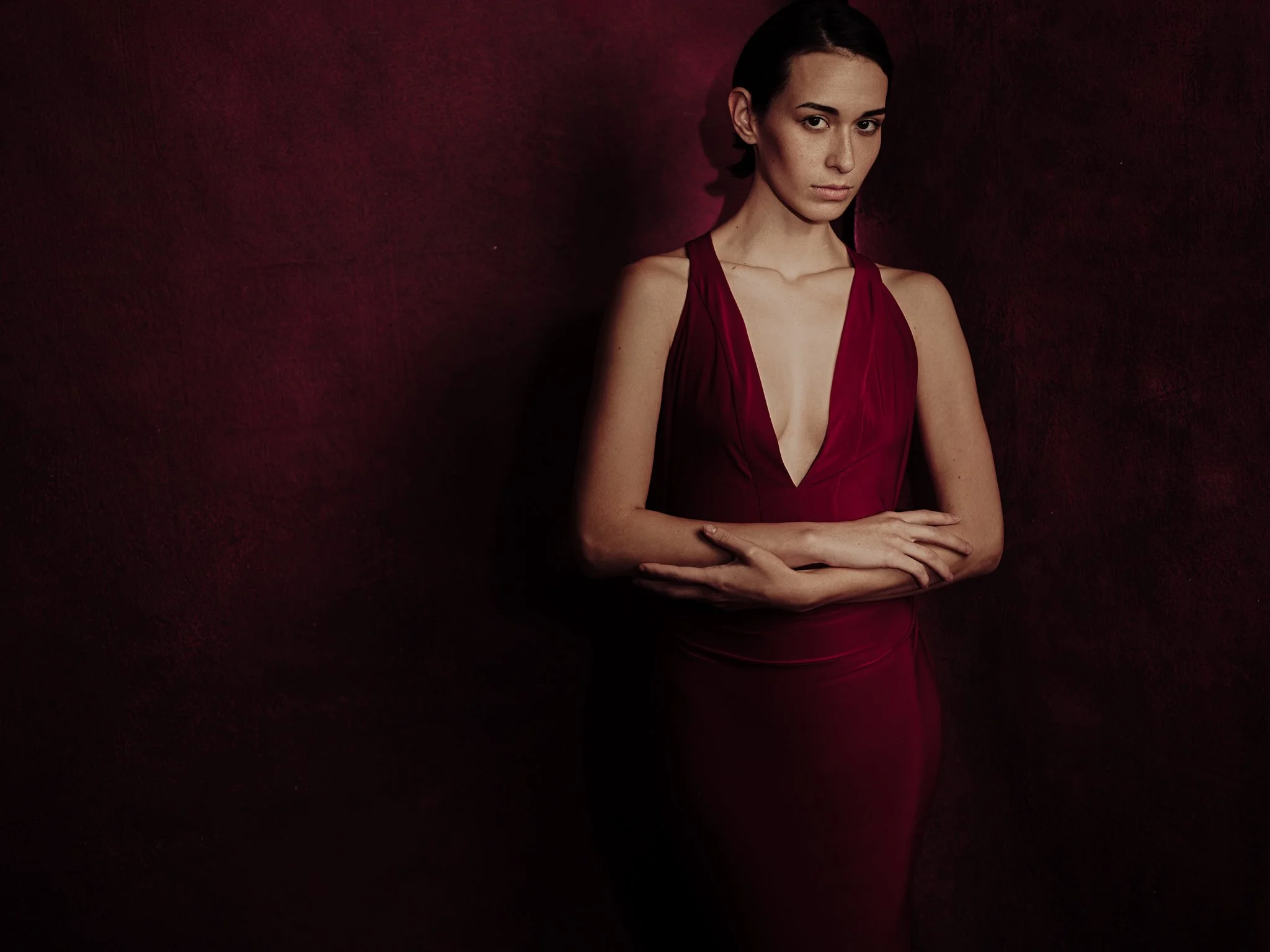 A woman in a deep red, sleeveless dress with a plunging neckline standing against a dark red background, with her arms crossed and looking at the camera with a serious expression.