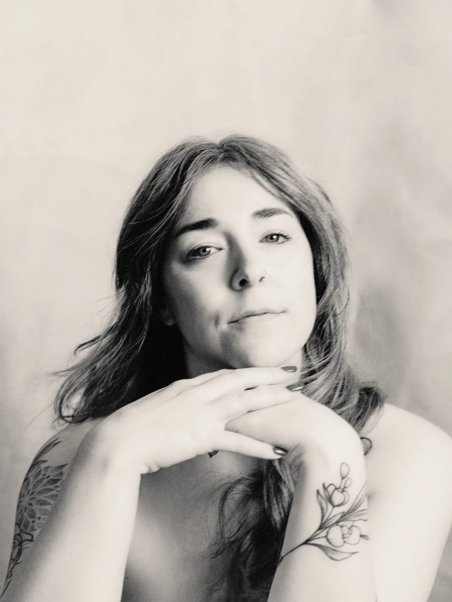 Black and white portrait of a young woman with long, wavy hair, resting her chin on her hands, tattoos on her arm, and looking directly at the camera.