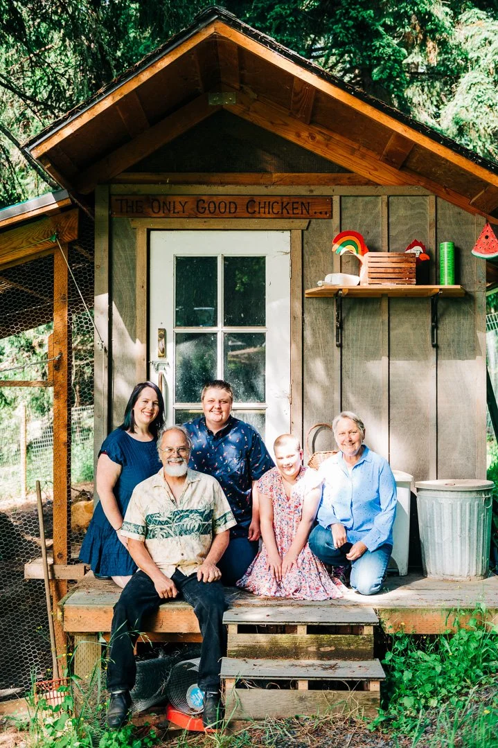 A group of five people posing in front of a small wooden chicken coop with a sign that reads "The Only Good Chicken..." surrounded by trees and greenery.