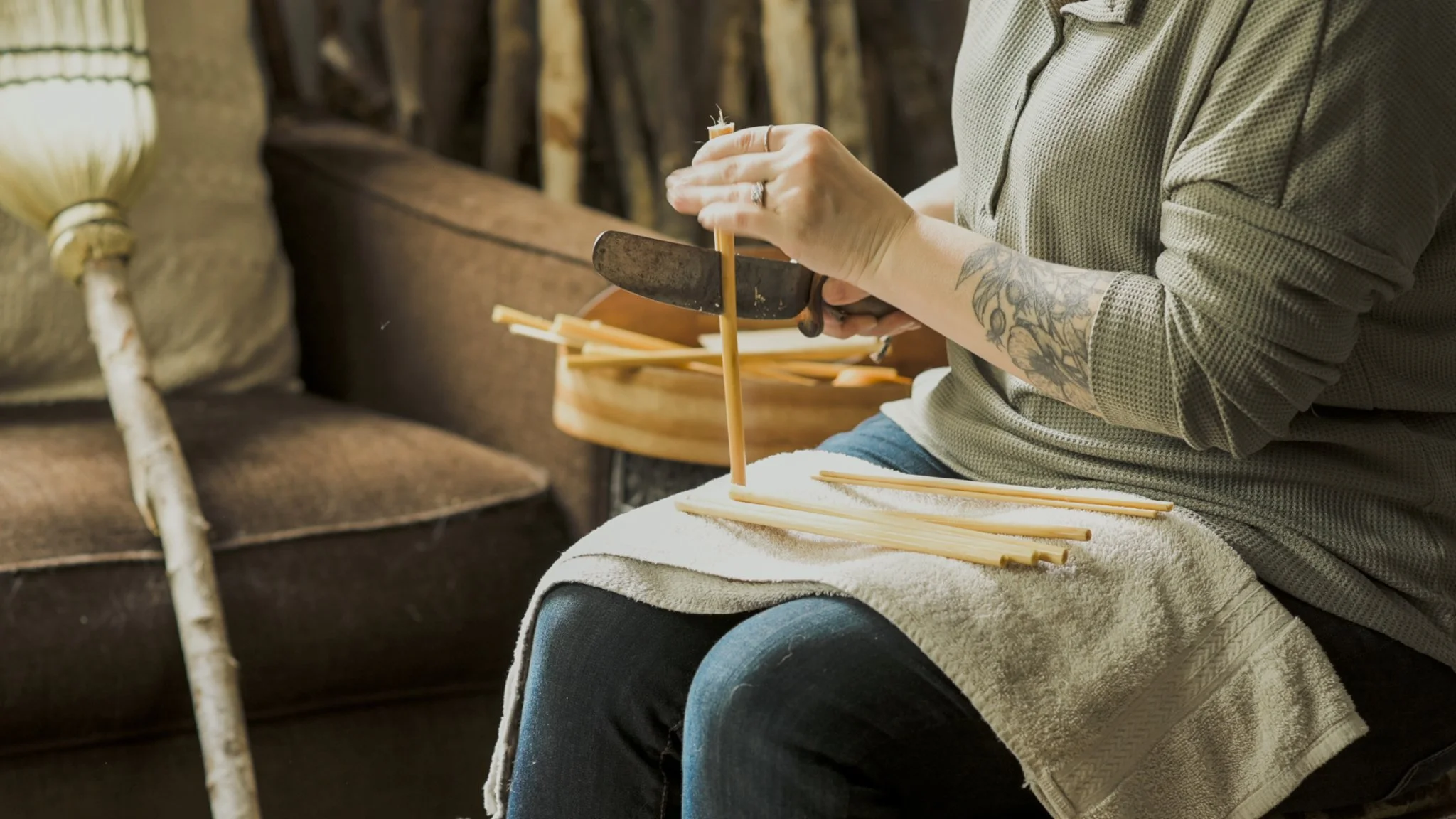 Person assembling bamboo chopsticks with a small knife, sitting on a towel-covered lap in a cozy, rustic setting.