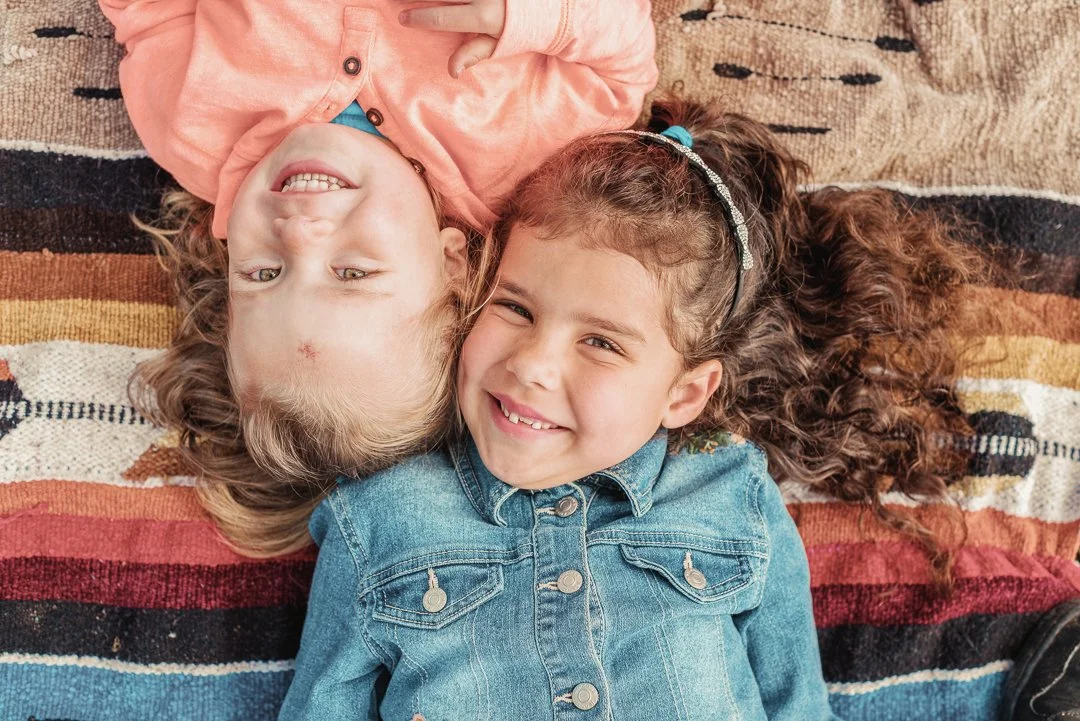 Two young girls lying on a colorful striped blanket, smiling at the camera. One girl has curly hair and a denim jacket, while the other has lighter hair and is wearing a pink jacket.