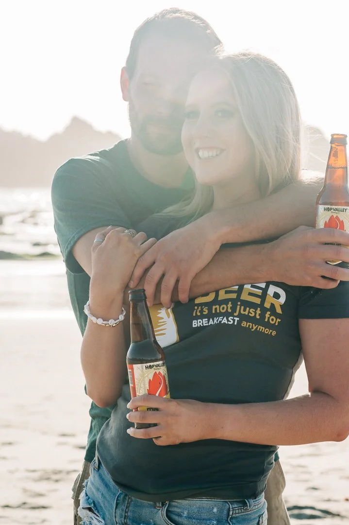 A couple hugging on the beach, both holding bottles of Beer. The woman is smiling and wearing a dark shirt with partially visible yellow text, while the man is behind her with his arms around her neck. The sun is bright, creating a backlit effect.