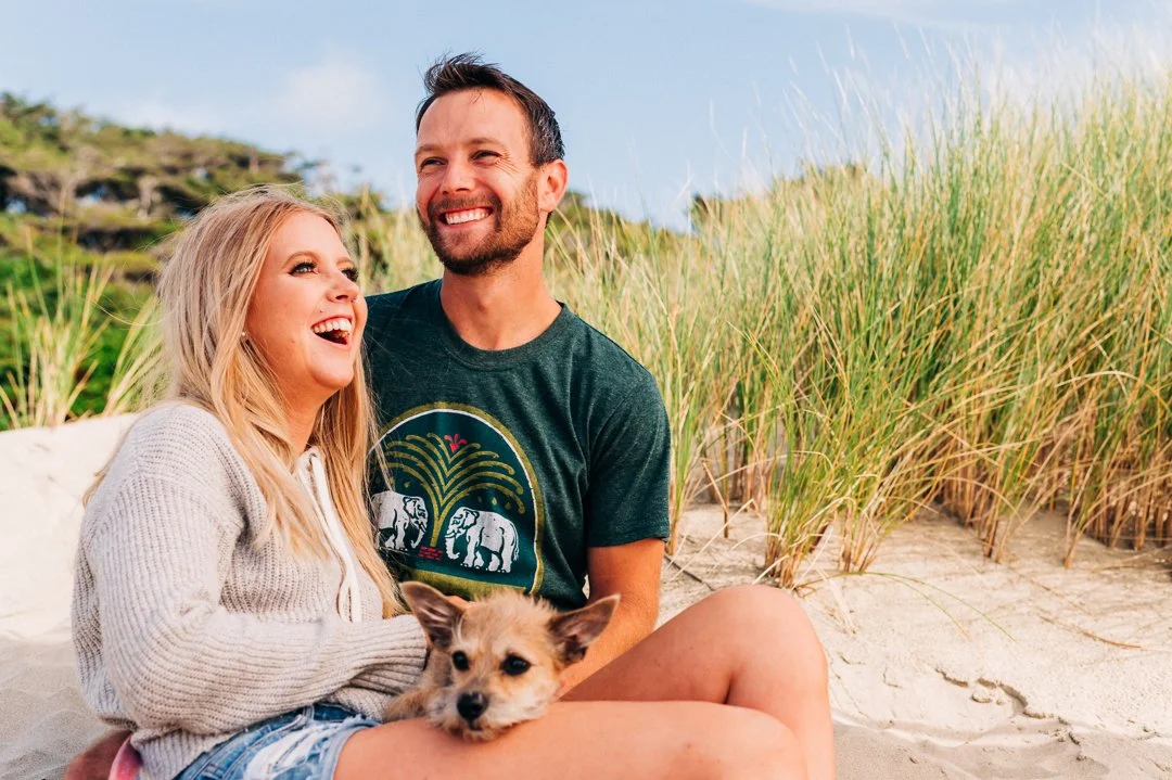 A happy couple sitting on a sandy beach with tall grass, smiling and laughing. The woman has long blonde hair and is holding a small dog. The man has short dark hair and a beard, wearing a dark t-shirt with a graphic design.