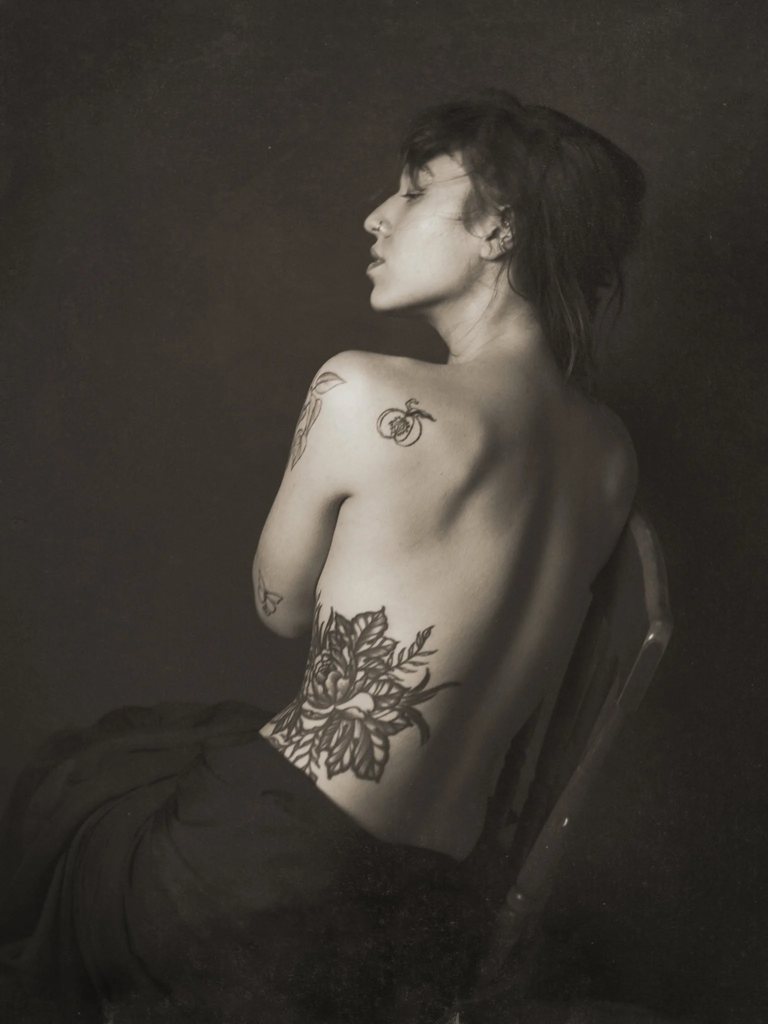 A black and white photo of a woman with tattoos on her shoulder and side, sitting sideways on a chair with her eyes closed and head tilted back, in a minimalist setting.